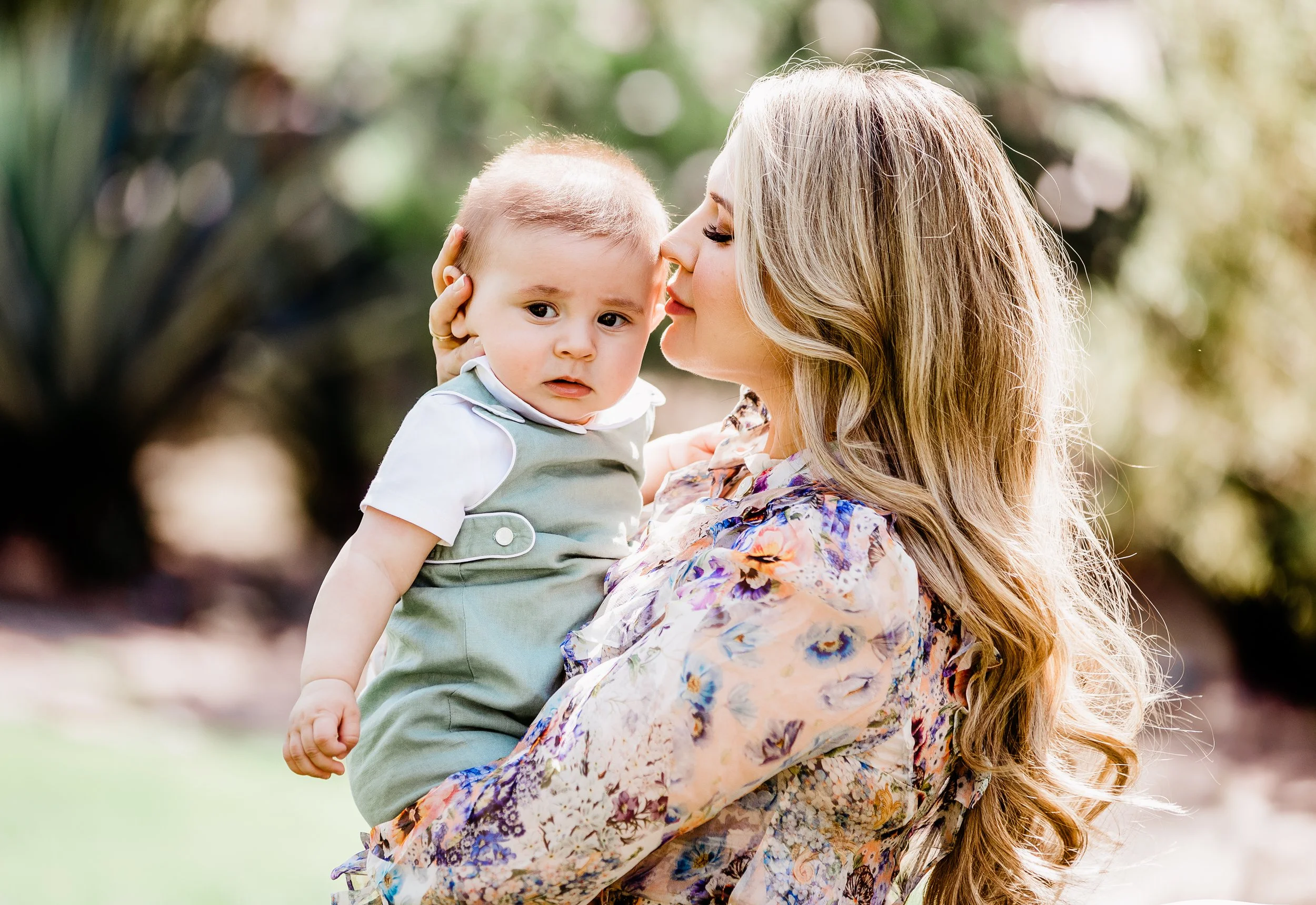 A woman with long wavy blonde hair holding a young child in an outdoor setting with greenery, the woman is kissing the child's cheek and the child has a concerned expression.