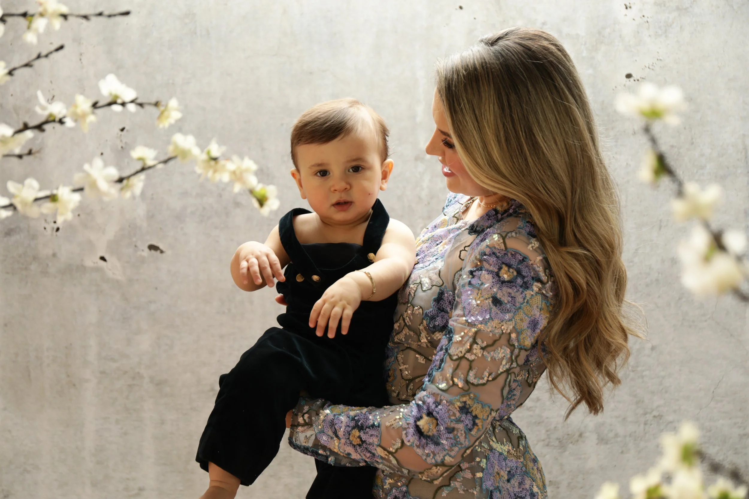 A woman holding a young child, both smiling, with white flowers in the foreground, and a plain gray background.
