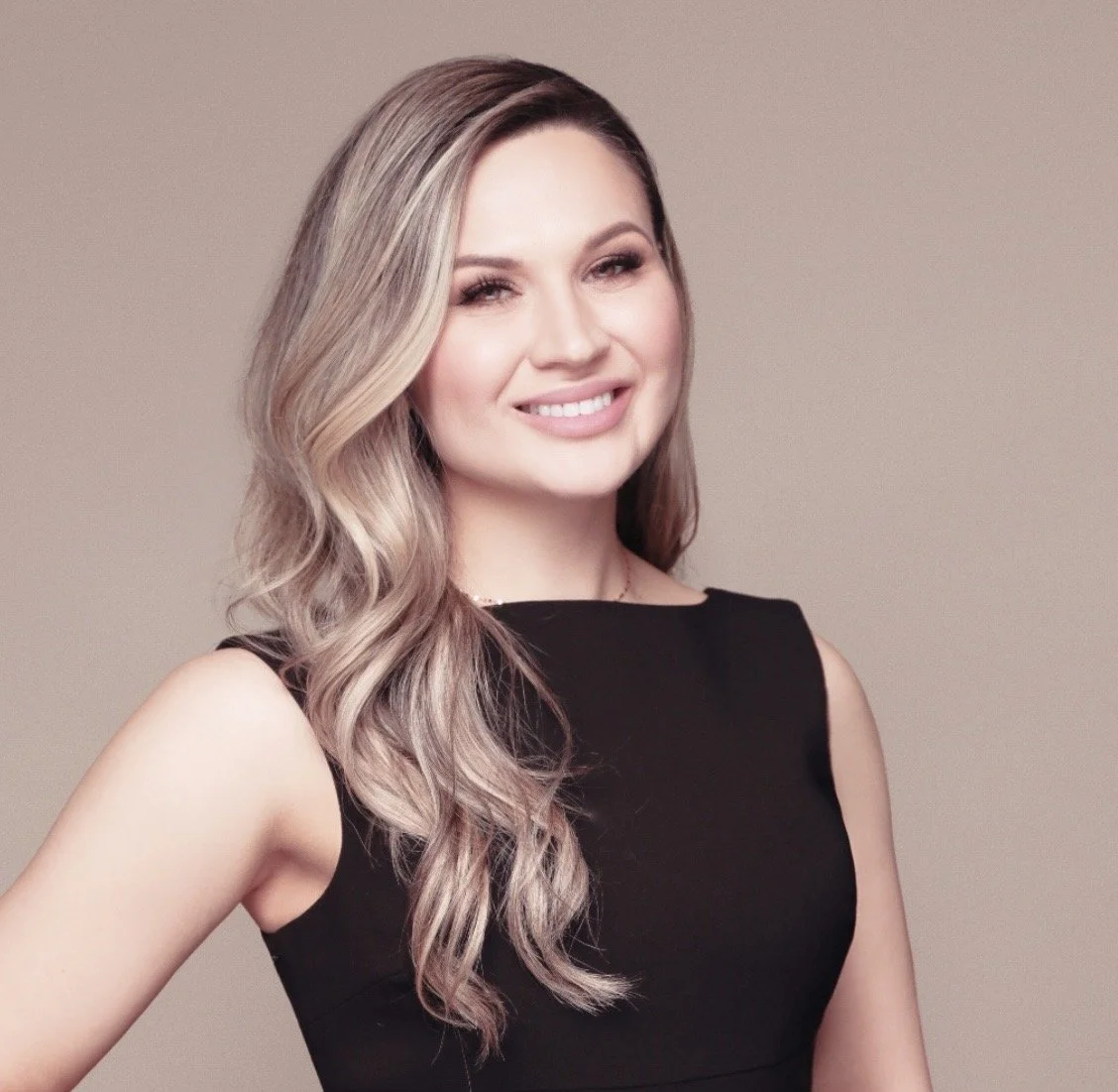 Dr. Nez, a pediatric dentist. with long, wavy blonde hair wearing a sleeveless black dress, smiling in front of a plain beige background.