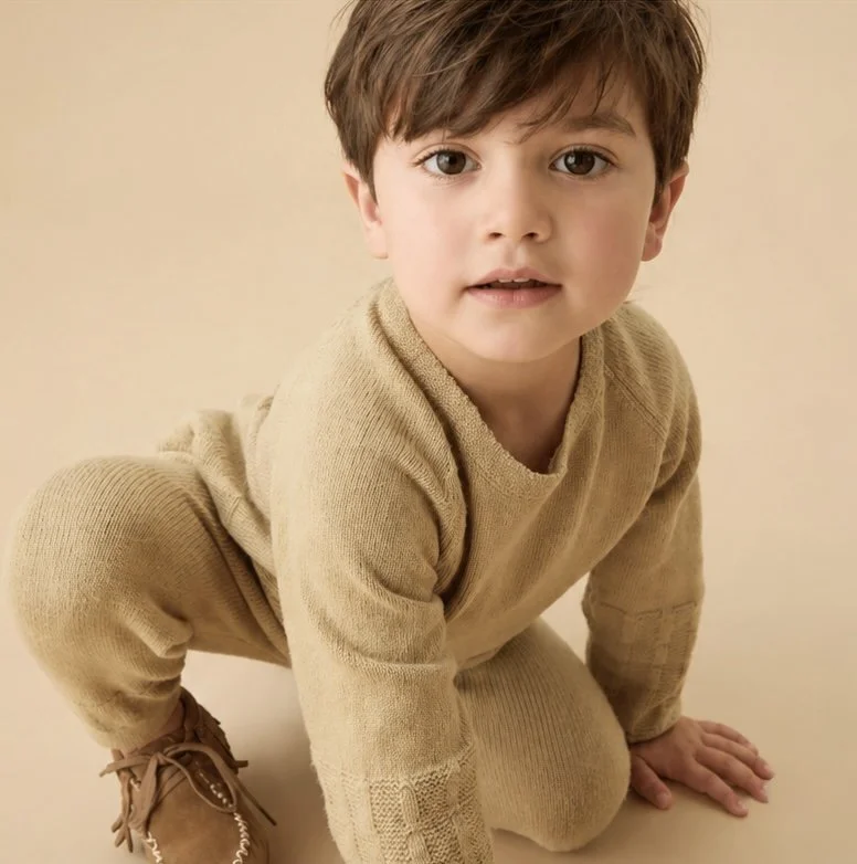 Quality sleep essential for growth showing a young child wearing a beige knit outfit and brown shoes kneels on a matching beige background, looking at the camera.