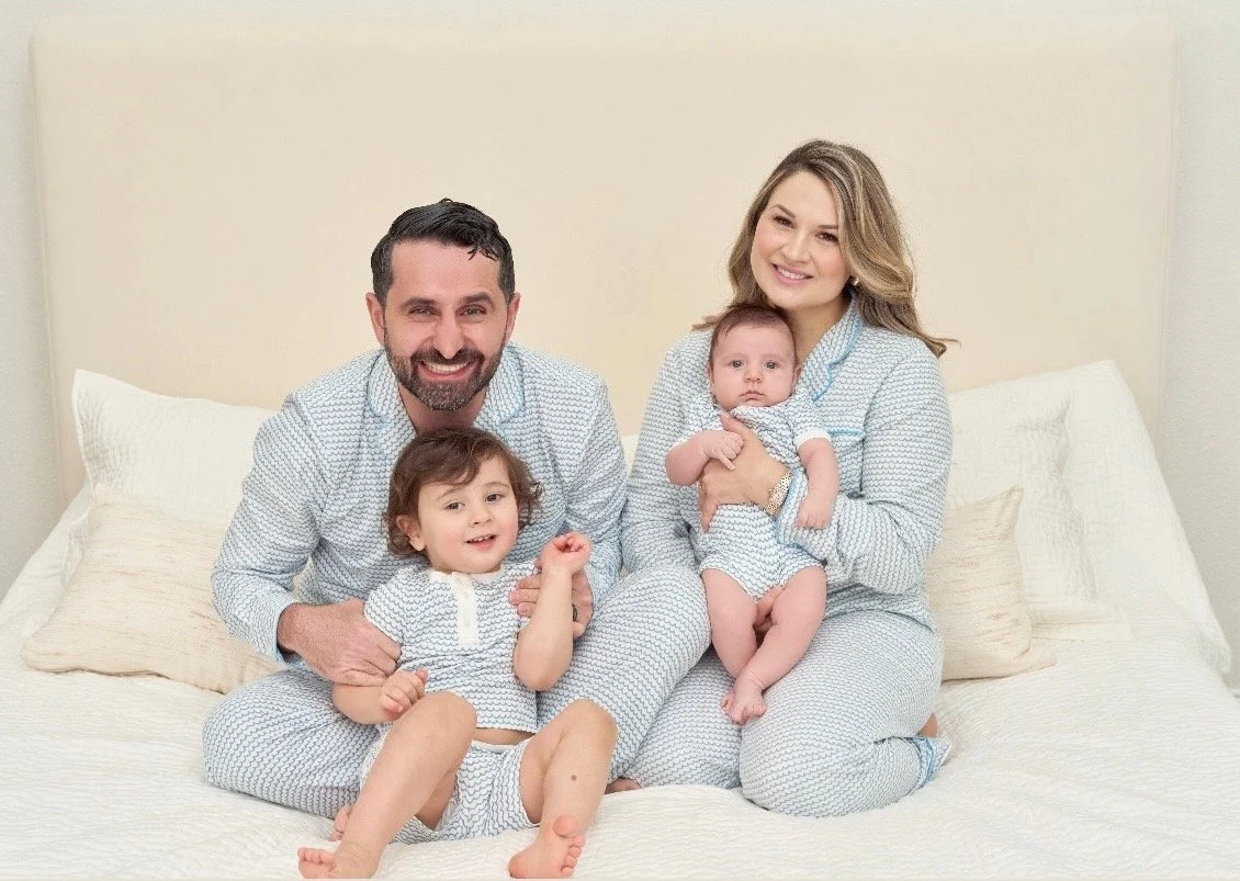 A family of four sitting on a bed, smiling at the camera. The father and a young girl are sitting in front, with the mother holding a baby girl. All are wearing matching pajamas with a light pattern.