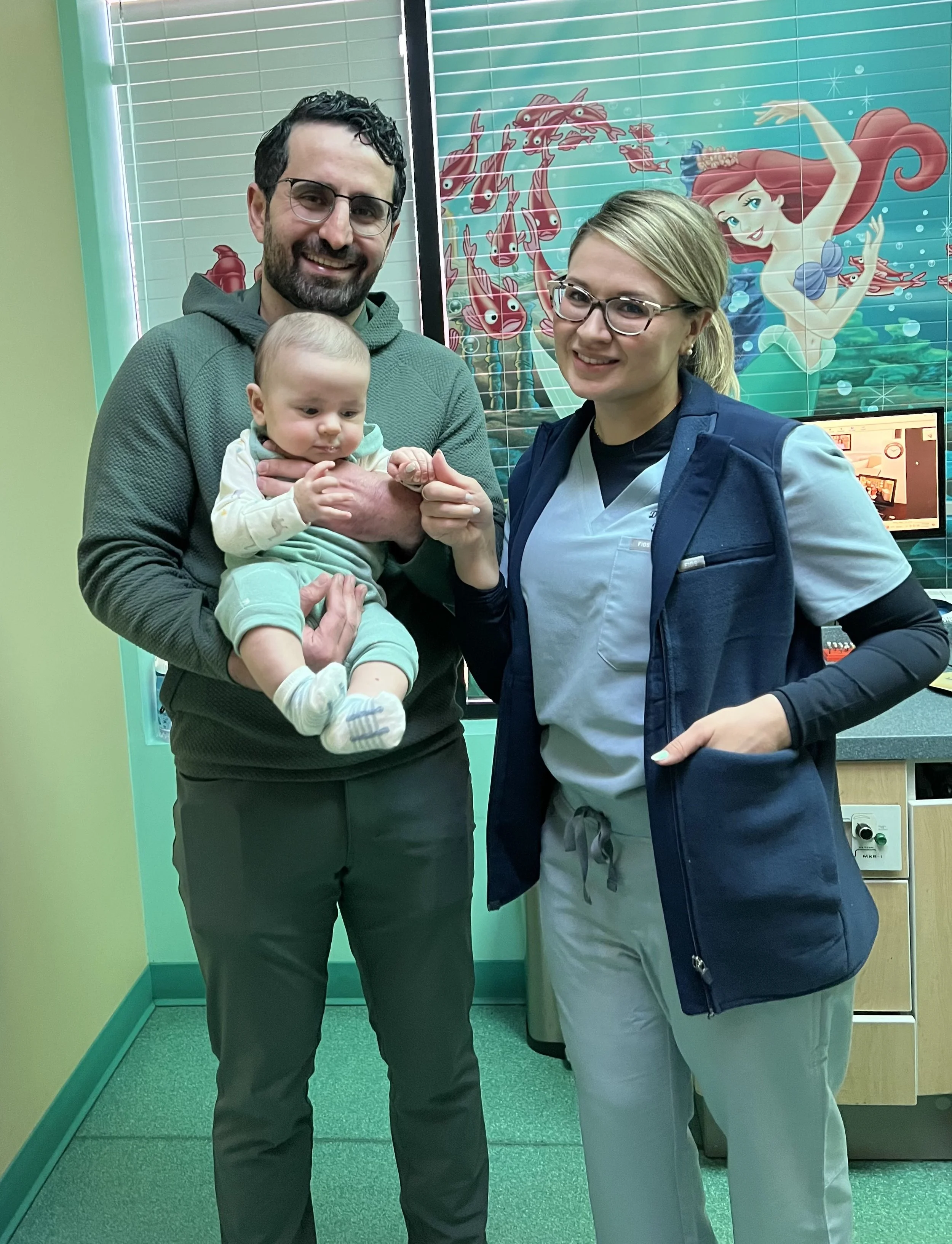 Man holding a baby in a clinic, with a female healthcare worker standing next to them. There is a colorful underwater-themed mural of Ariel from The Little Mermaid in the background.