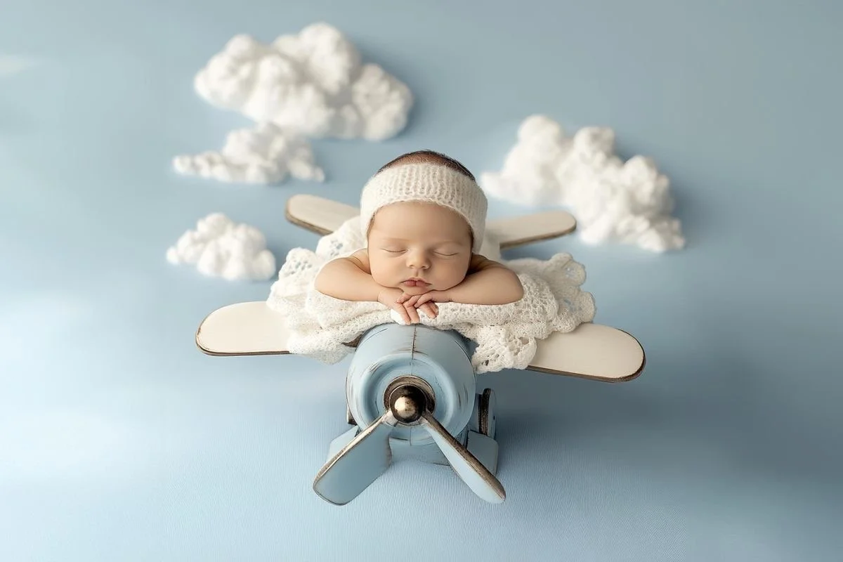 A sleeping baby with a knitted hat, resting on a toy airplane with clouds and a blue sky background.