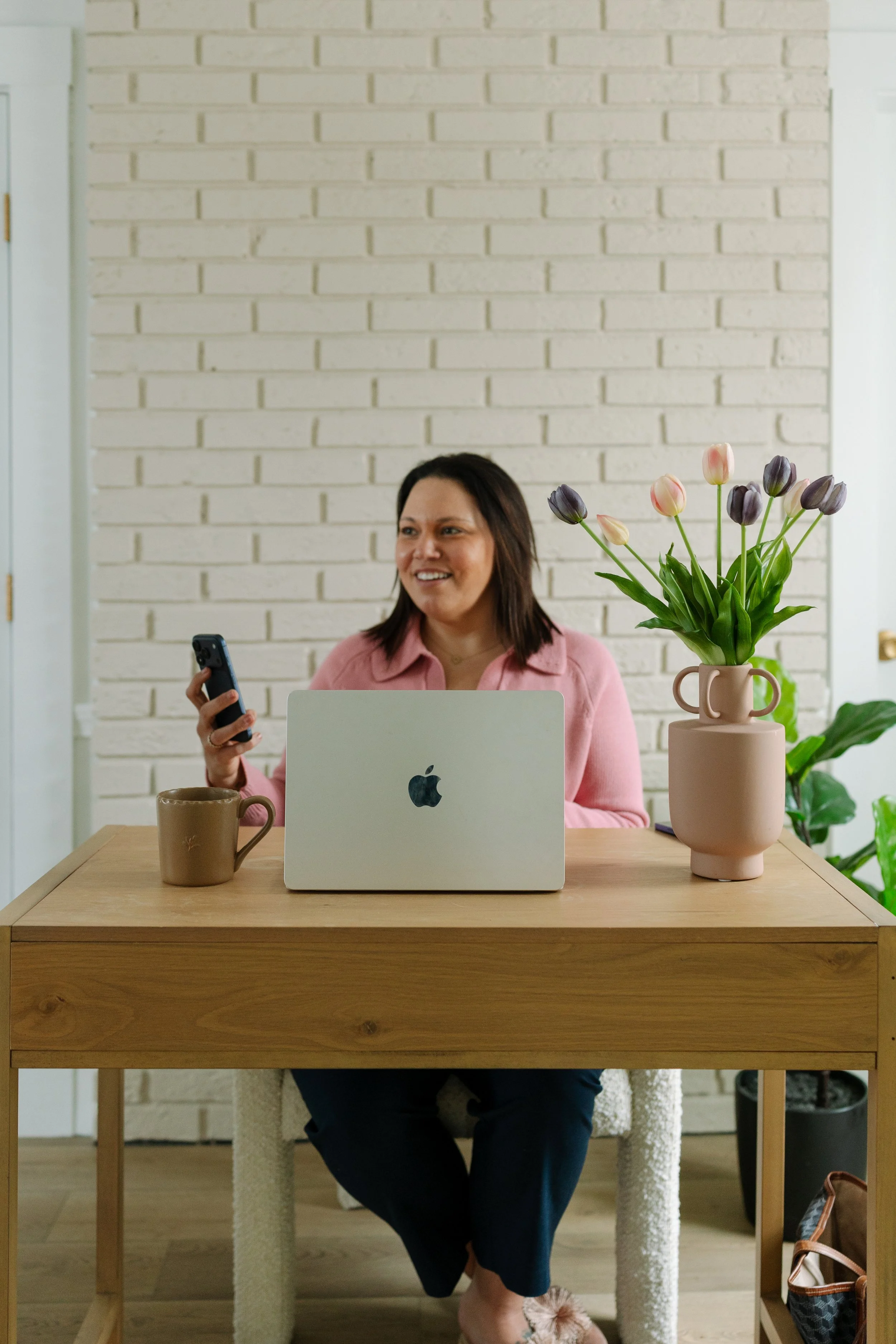 A woman with brown hair, wearing a pink shirt, sits at a wooden desk with an Apple laptop, a beige ceramic mug, and a pink vase with tulips. She is holding a smartphone and smiling, with a white brick wall and green plant in the background.
