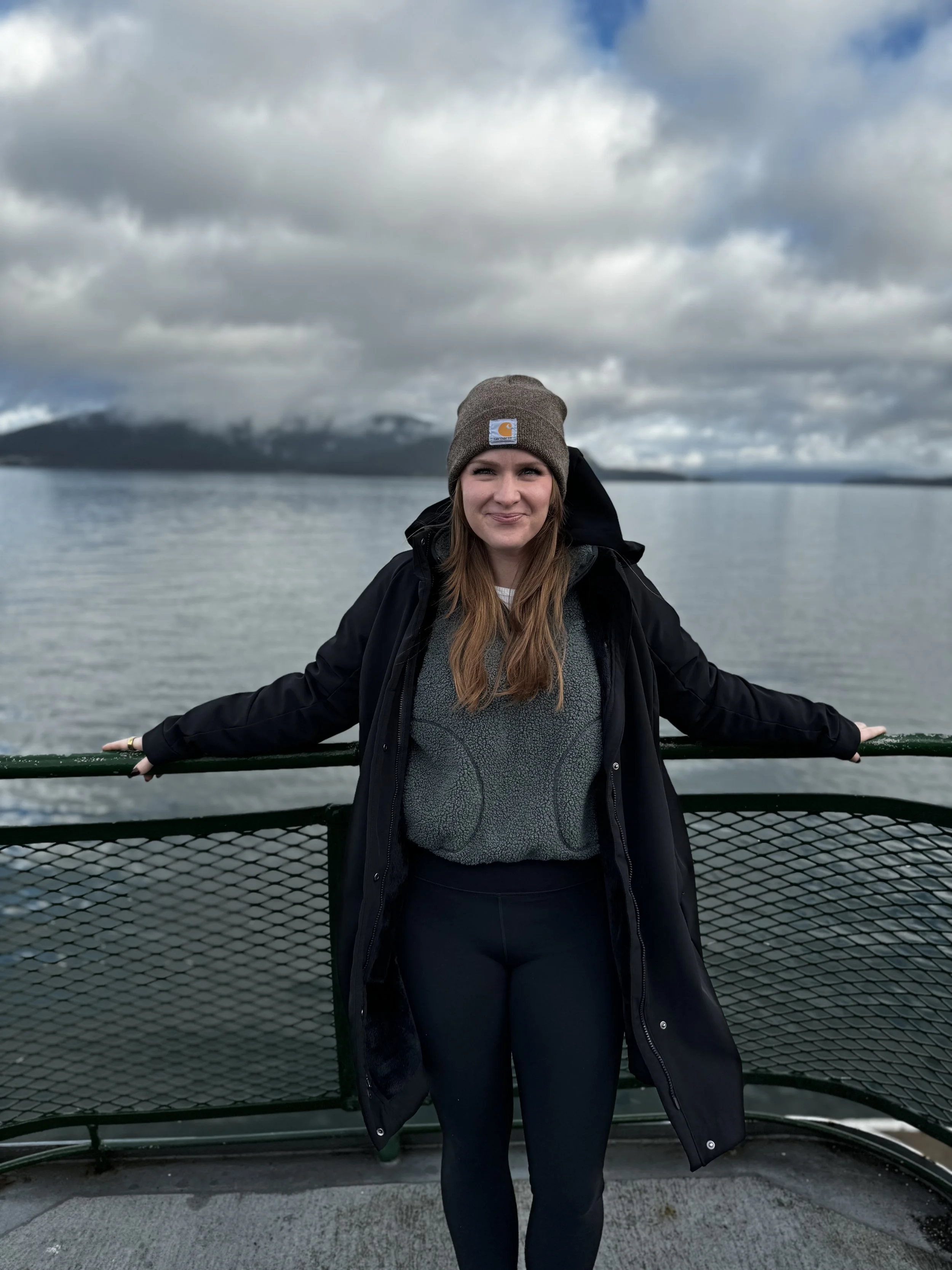 A young woman standing on a boat deck with her arms extended, wearing a gray beanie, black jacket, and black leggings, with a body of water and cloudy sky in the background.