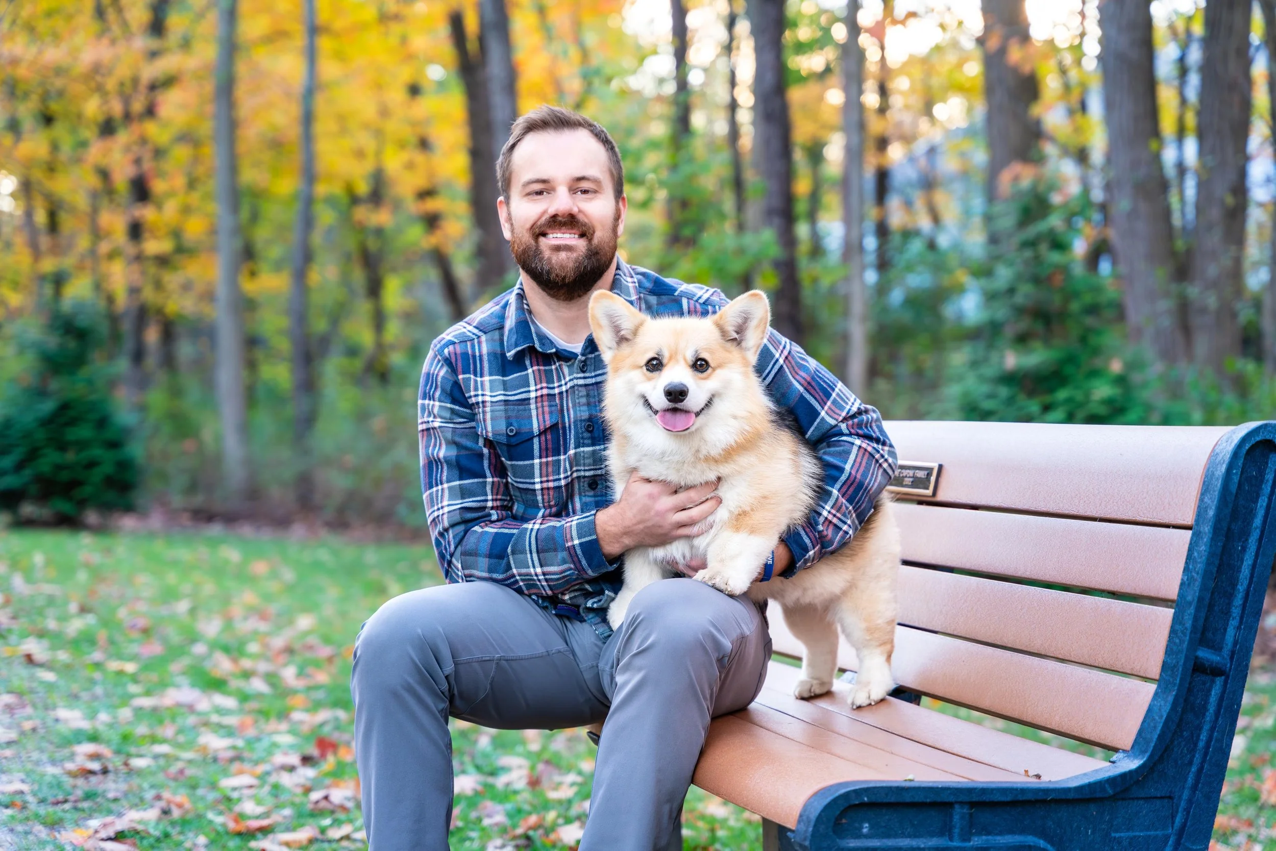 A man with a beard and a woman in a plaid shirt sitting on a park bench, holding a smiling corgi dog, with fall trees and colorful leaves in the background.