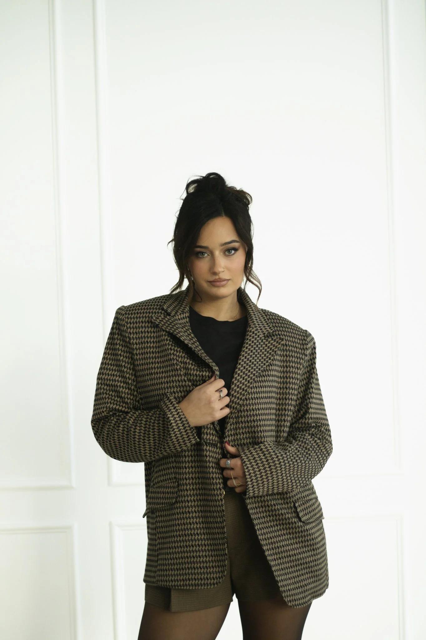 A woman with dark hair in a messy bun stands in front of a plain white wall, wearing a houndstooth blazer, a black top, and brown shorts with black tights.