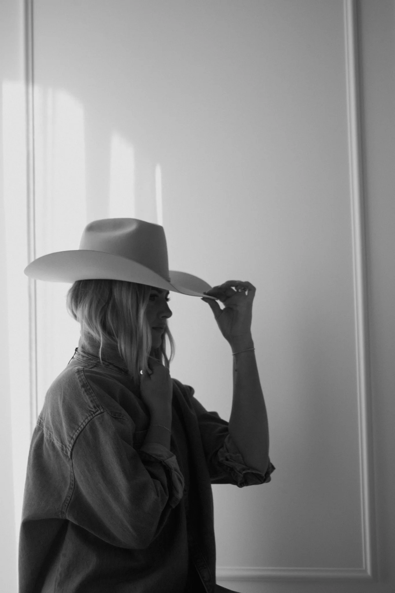 A woman with wavy hair wearing a cowboy hat and a denim jacket, standing against a plain wall, adjusting her hat.