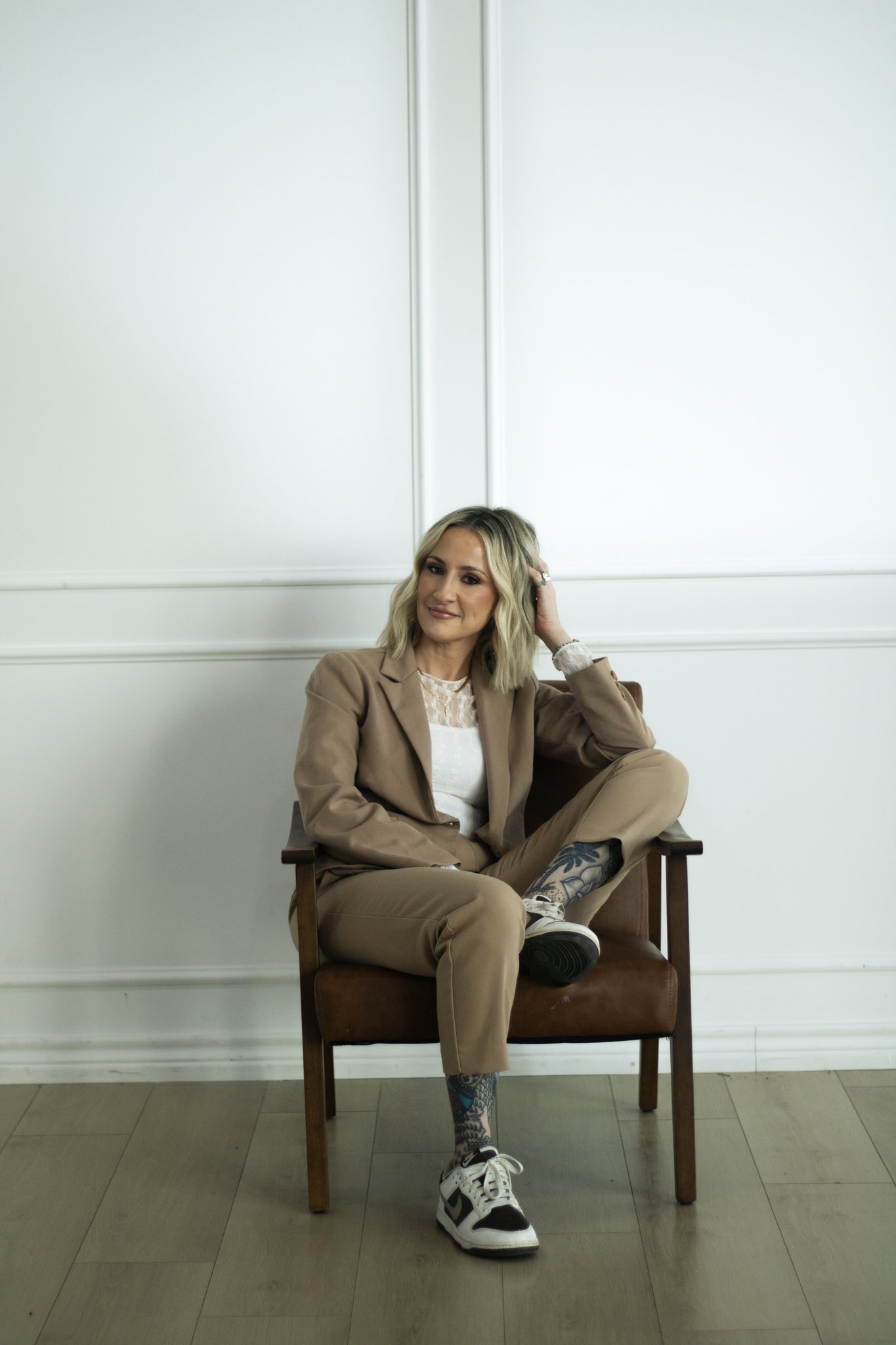 A woman with blonde hair, wearing a tan blazer and tan pants, sitting on a wooden chair against a plain white wall. She is smiling, resting her head on her left hand, and has one leg crossed over the other, revealing patterned socks and white sneakers.