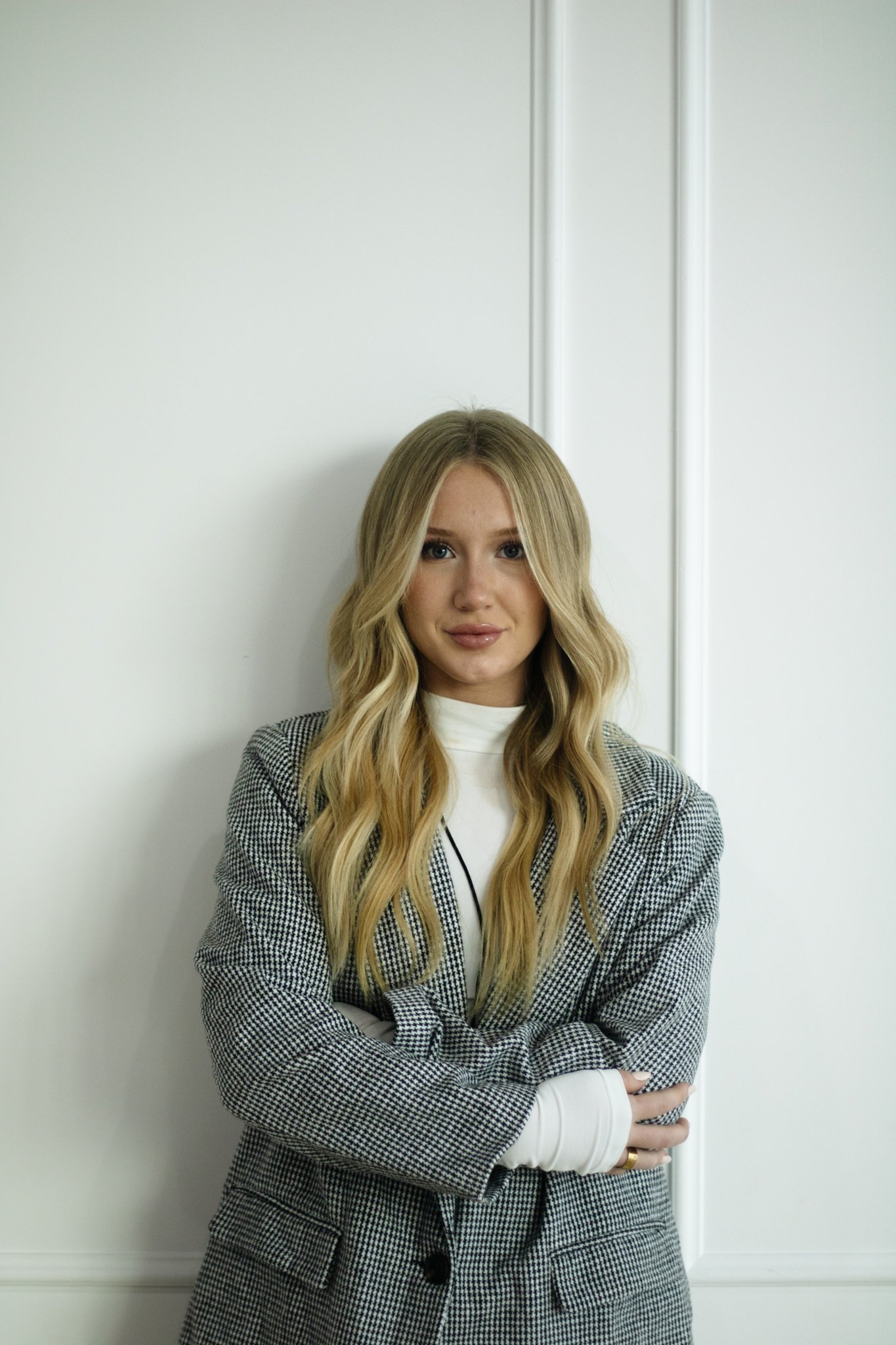 A young woman with long, wavy blonde hair standing against a white wall, wearing a black-and-white checkered blazer and a white turtleneck shirt.