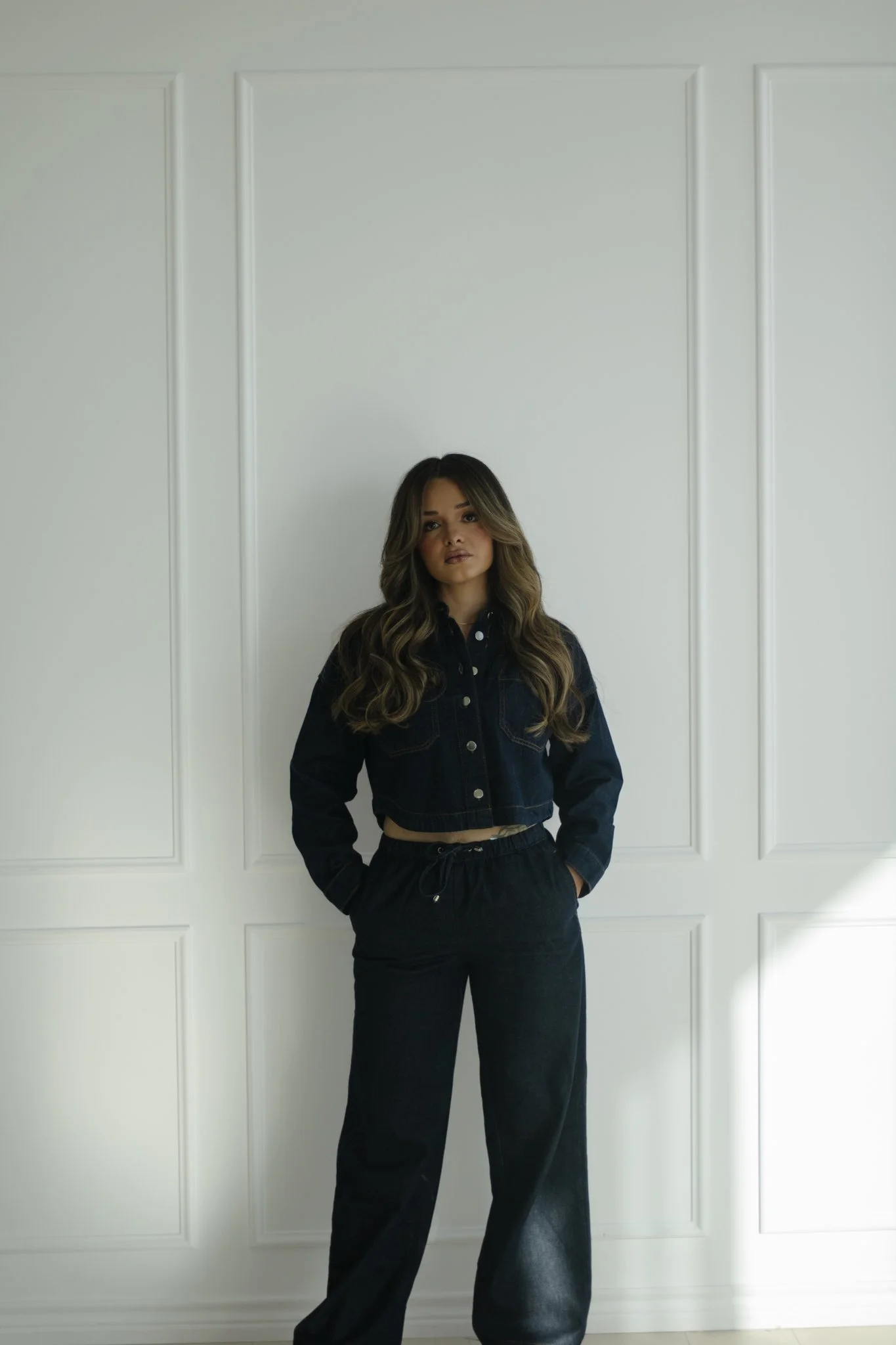 Young woman standing in front of a white wall with decorative molding, wearing a black denim jacket and black pants, with her hands in her pockets.