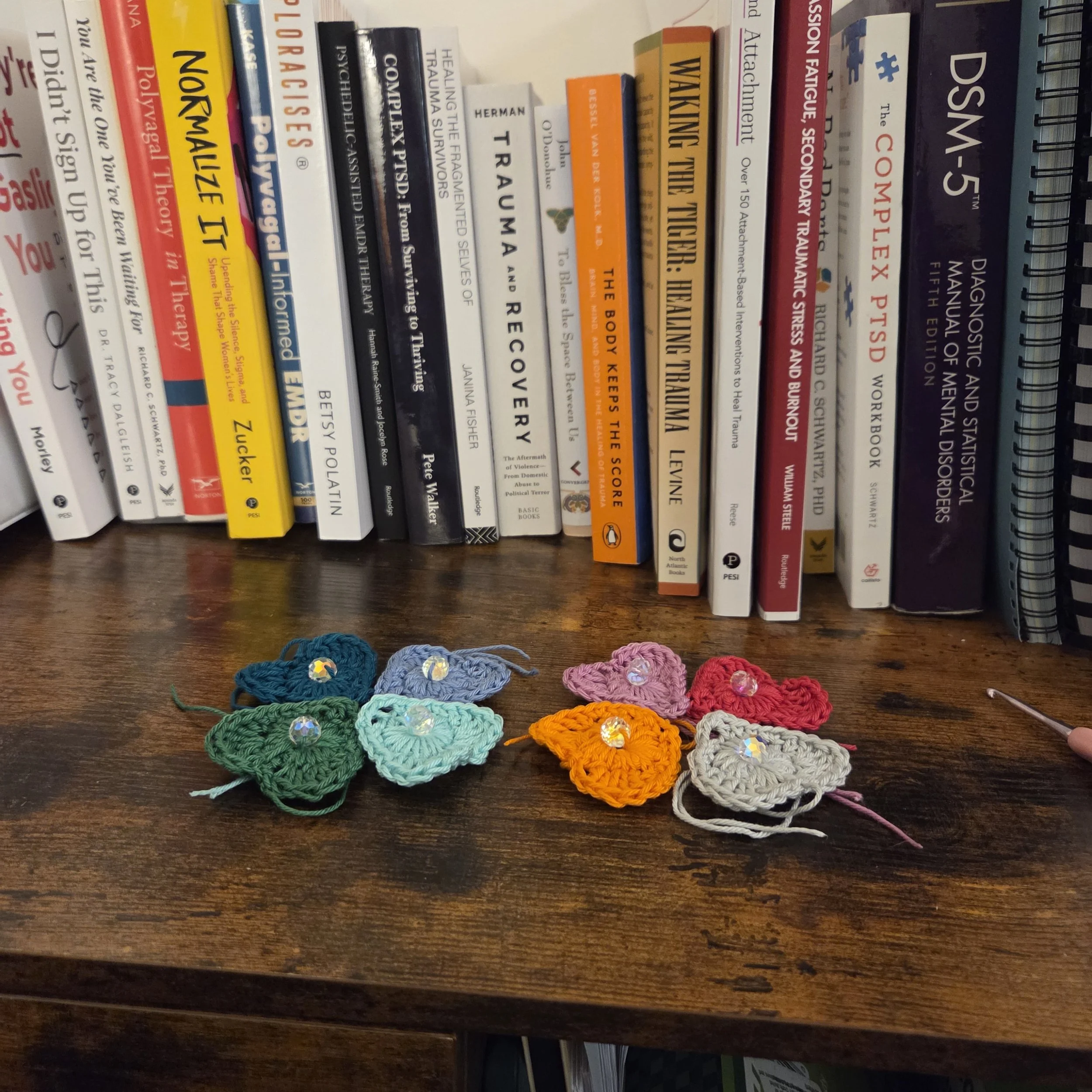 Colorful crocheted hearts with sparkly embellishments arranged on a wooden surface in front of a row of books on a bookshelf.