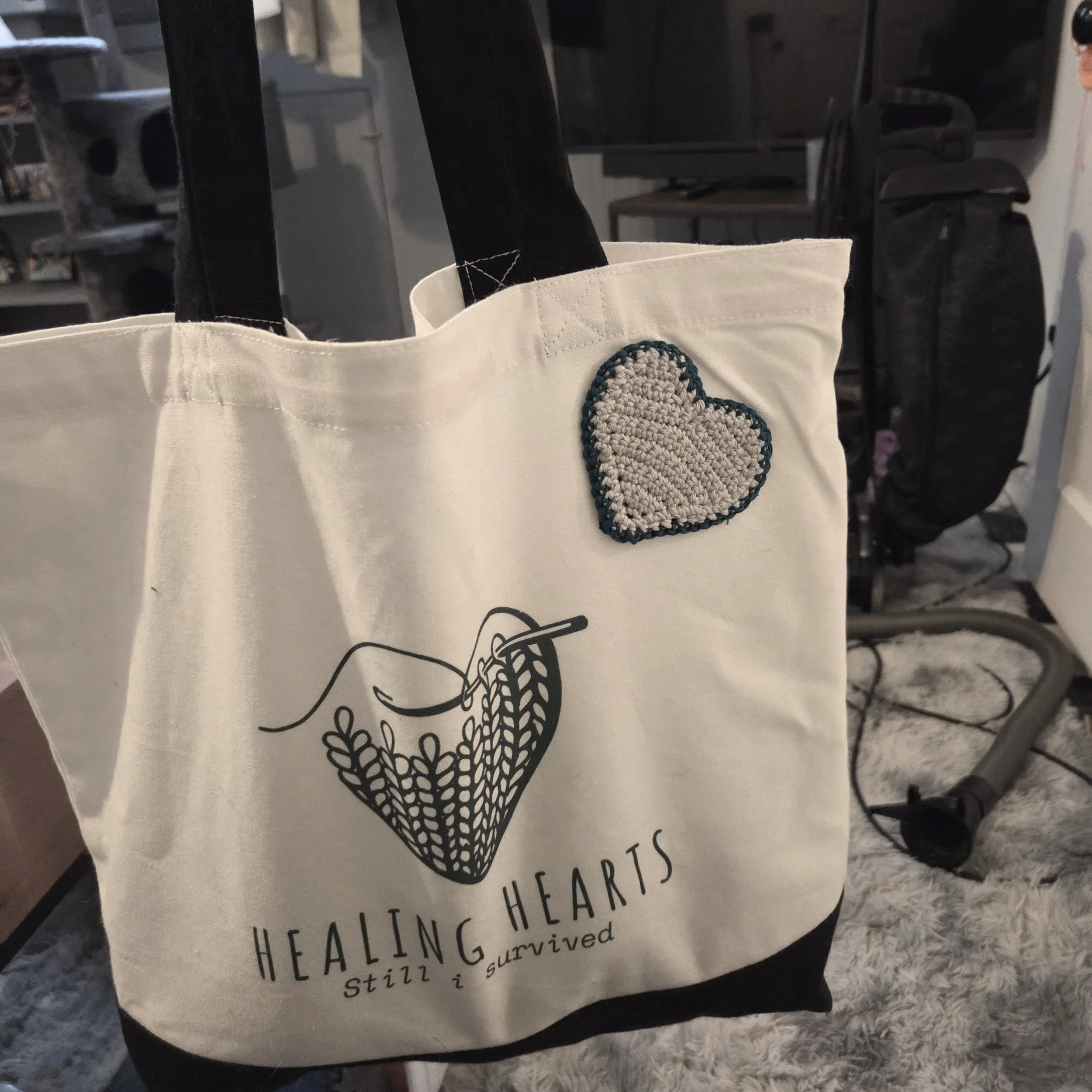 A white tote bag with black straps featuring a drawing of a knitting needle and heart shape, with the words 'Healing Hearts Still Survived' and a small heart embroidered on the top right corner.