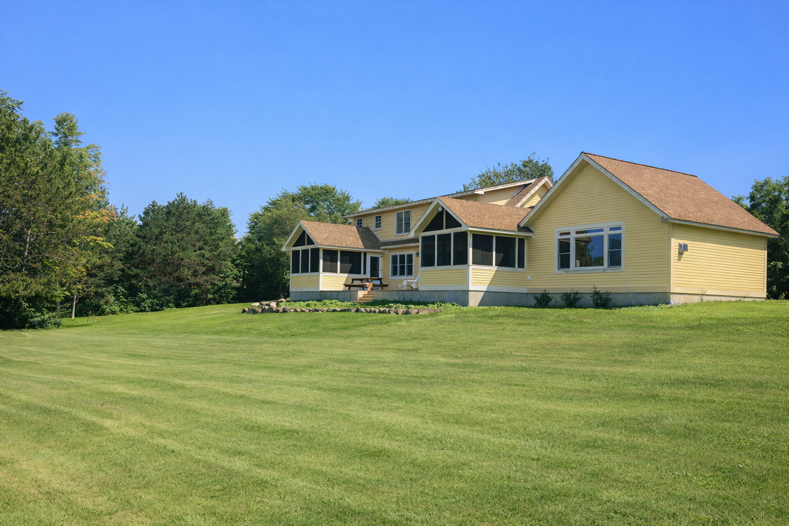 A yellow house with a porch set on a grassy lawn, surrounded by trees under a clear blue sky.