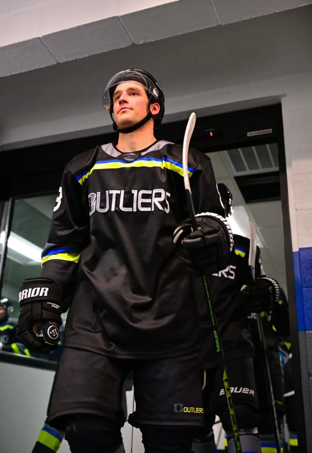 Hockey player in black and blue jersey holding a hockey stick, standing with other players behind in a locker room.
