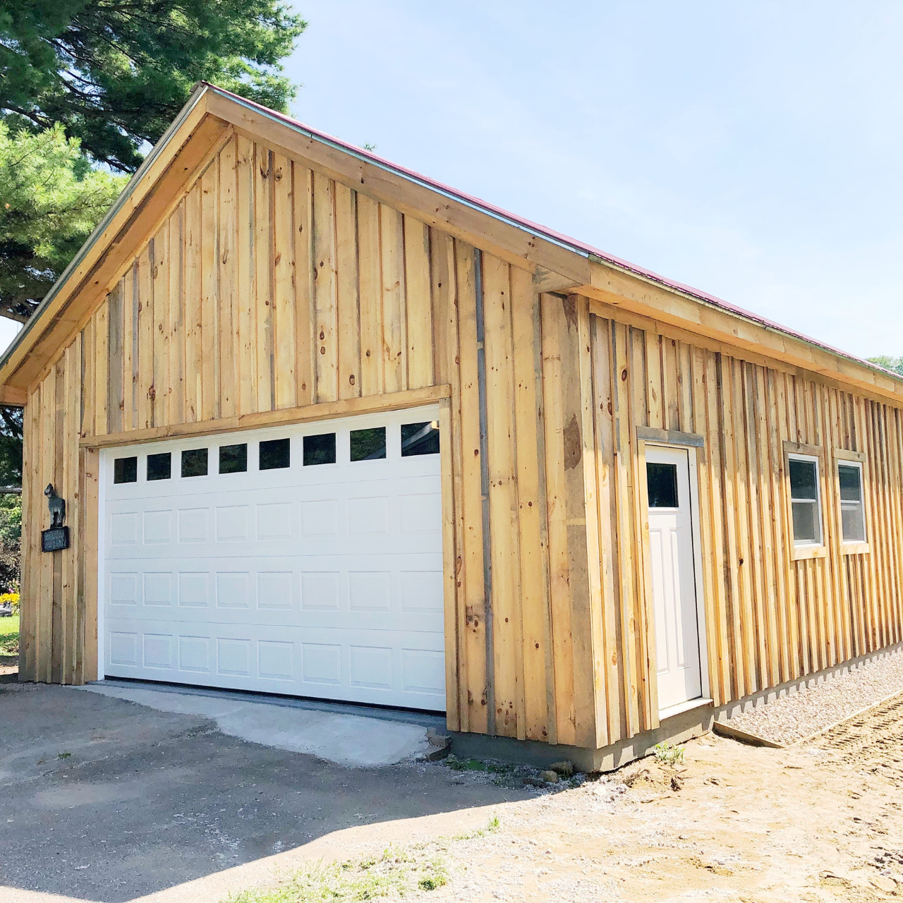 A newly built wooden garage with a white sectional roll-up door, a side door, and two small windows, surrounded by a gravel driveway and some trees.
