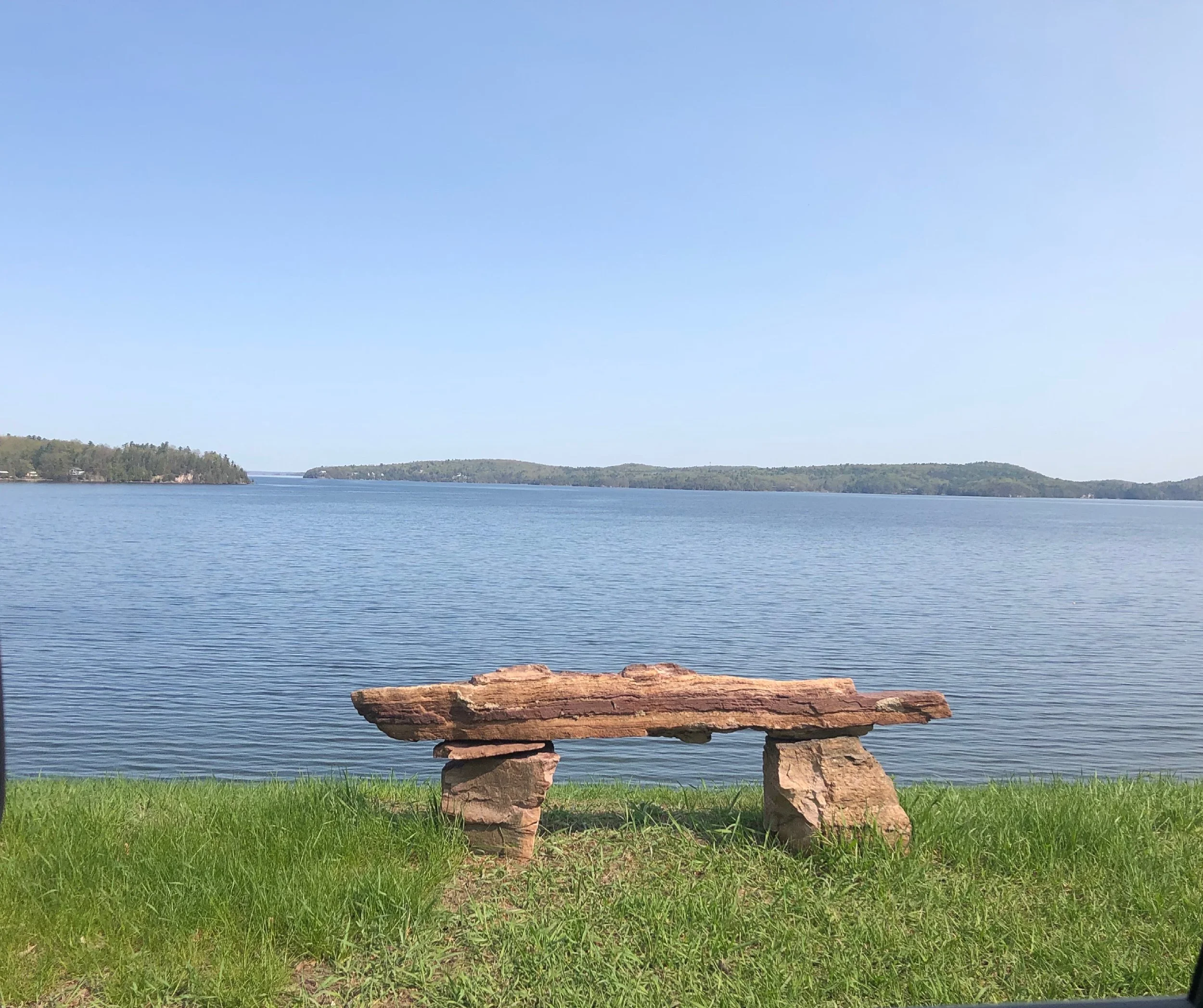 A rustic wooden bench made of uneven planks and rocks, positioned on lush green grass, overlooking a peaceful lake with small islands and distant tree-covered land under a clear blue sky.