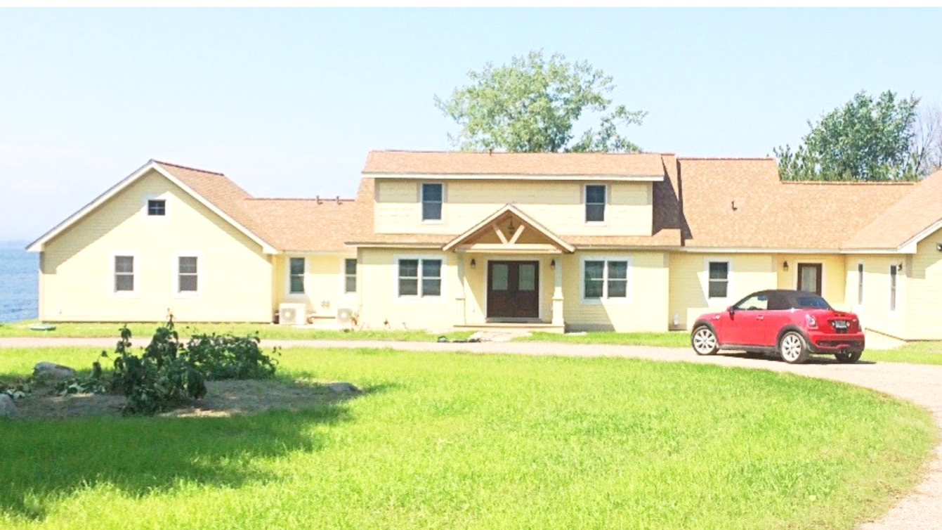 Yellow multi-story house with brown roof, black double front door, and a red car parked on driveway in front, with green lawn and trees in background.