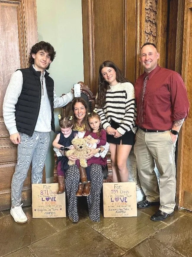 Family of five celebrating a pregnancy announcement, with signs indicating days until the baby's due date, gathering around a woman seated in a wooden chair holding a teddy bear and a child, in a room with wooden paneling.
