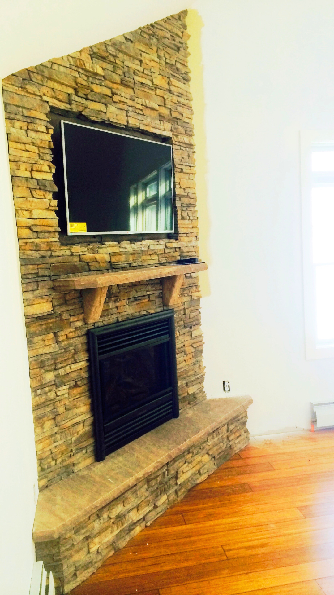Living room with a stone fireplace, a wooden mantel, and a flat-screen TV mounted above the mantel, with a window to the right.