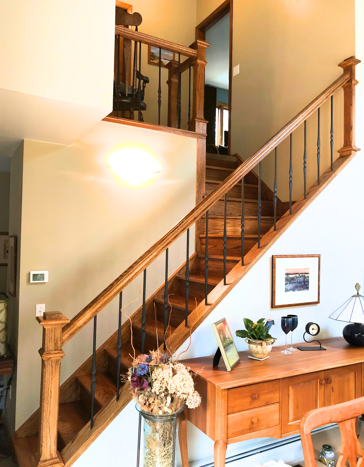 Interior of a house with a wooden staircase, a sideboard with decorative items, and wall art.