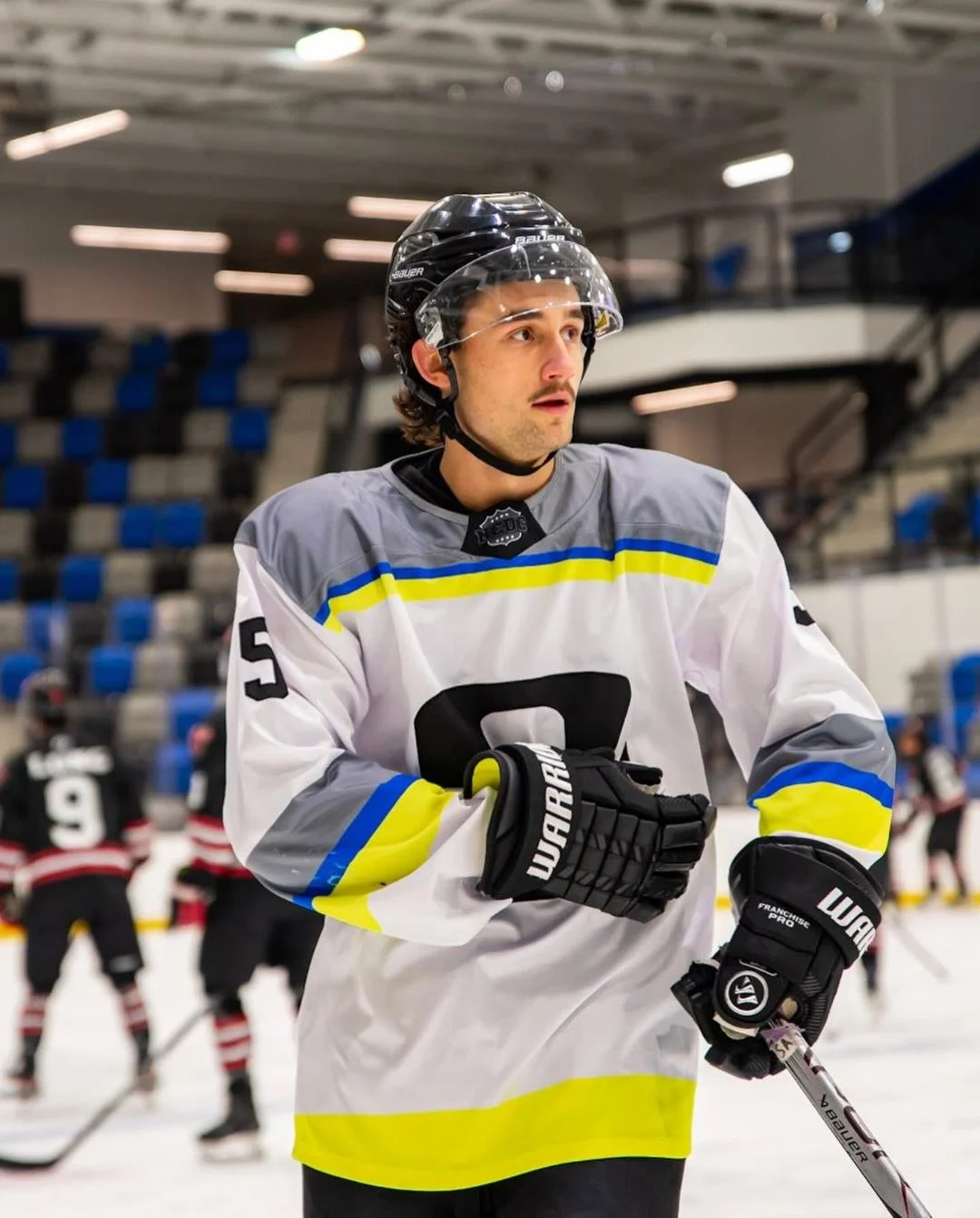 Ice hockey player in a gray and white jersey with yellow and blue accents, wearing a black helmet with a clear visor, standing on an ice rink.
