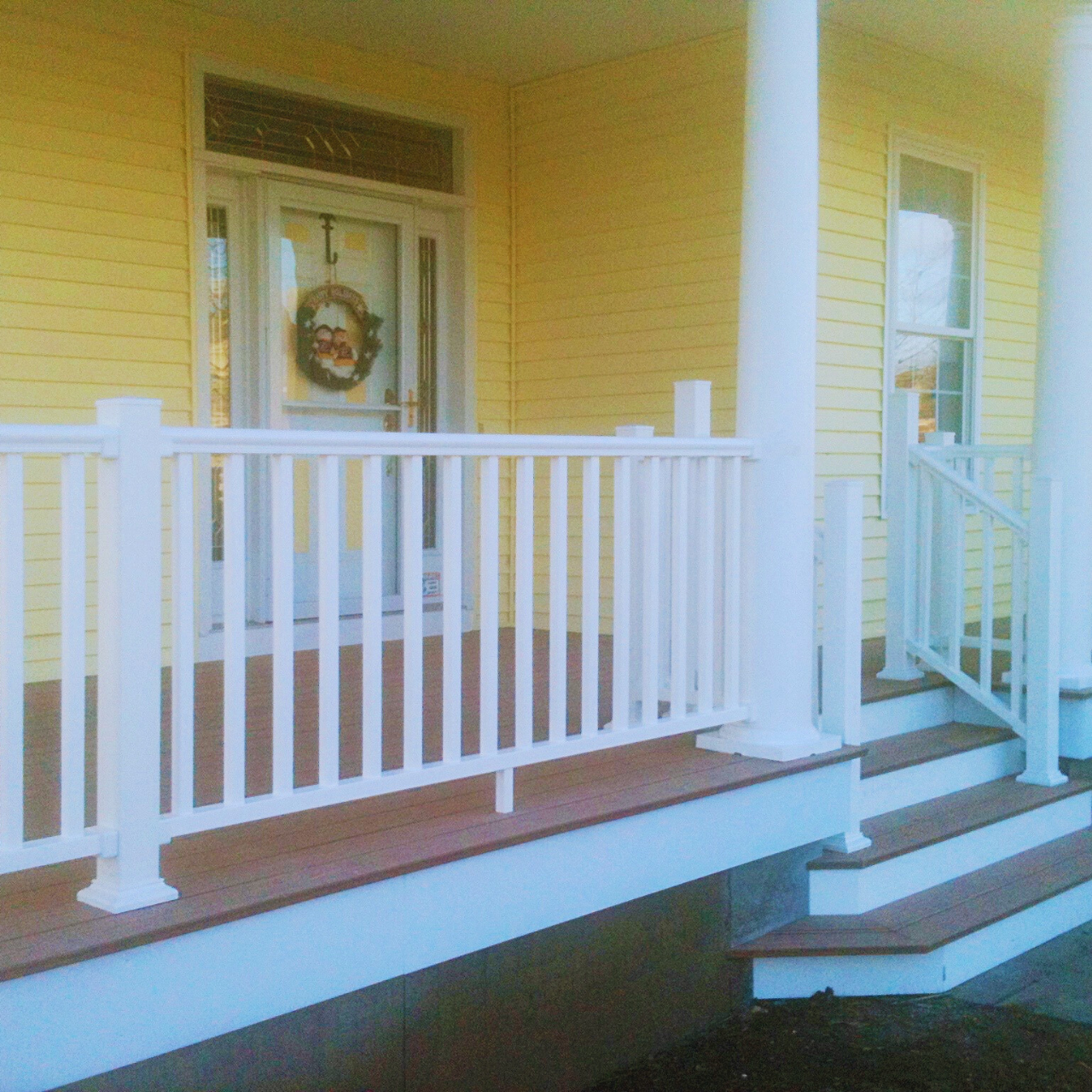 Close-up of a yellow house porch with white railing and steps, front door with holiday wreath, and a window to the right.