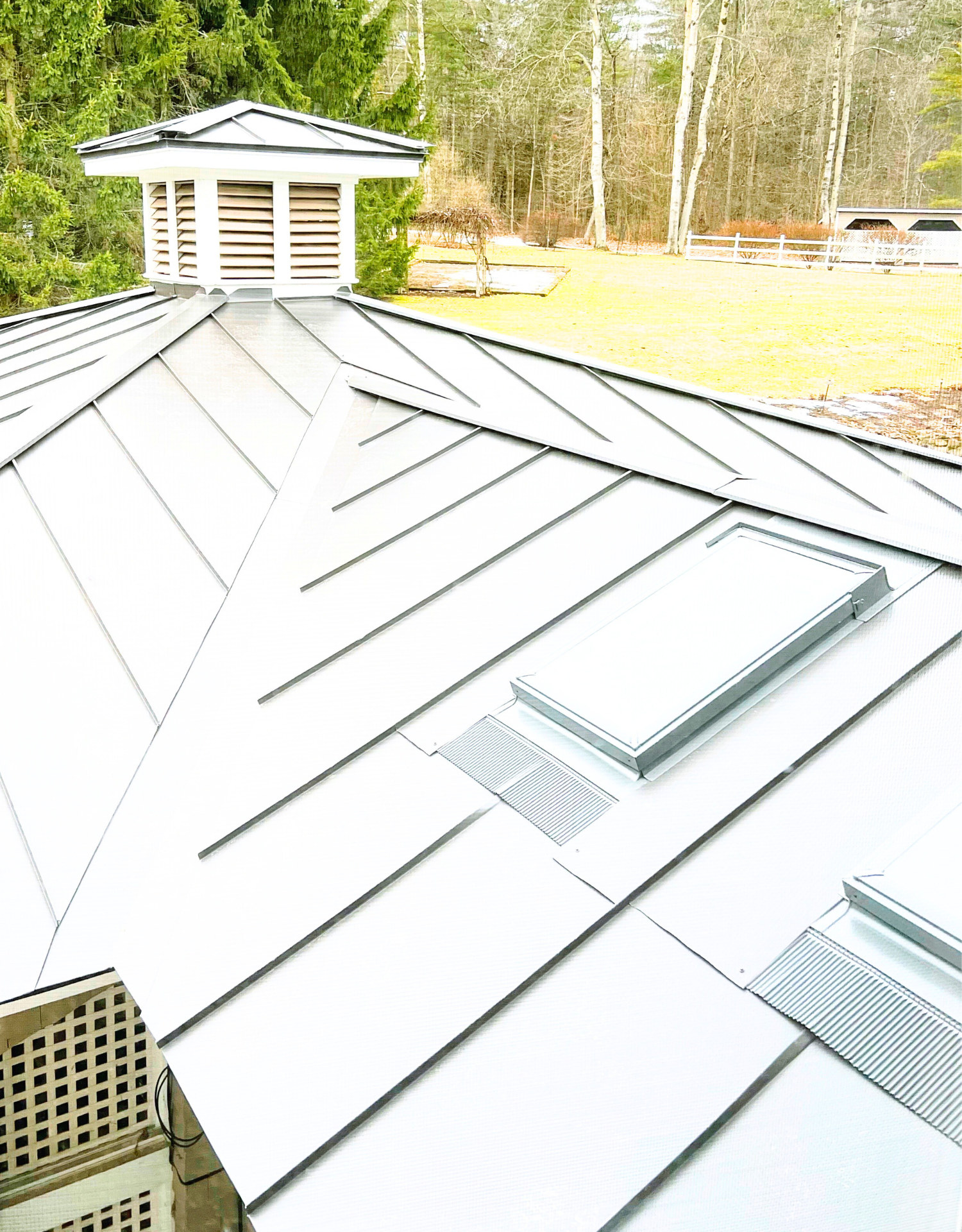 A close-up view of a metal roof with two skylights, surrounded by a rural landscape with trees, grass, and a fence.