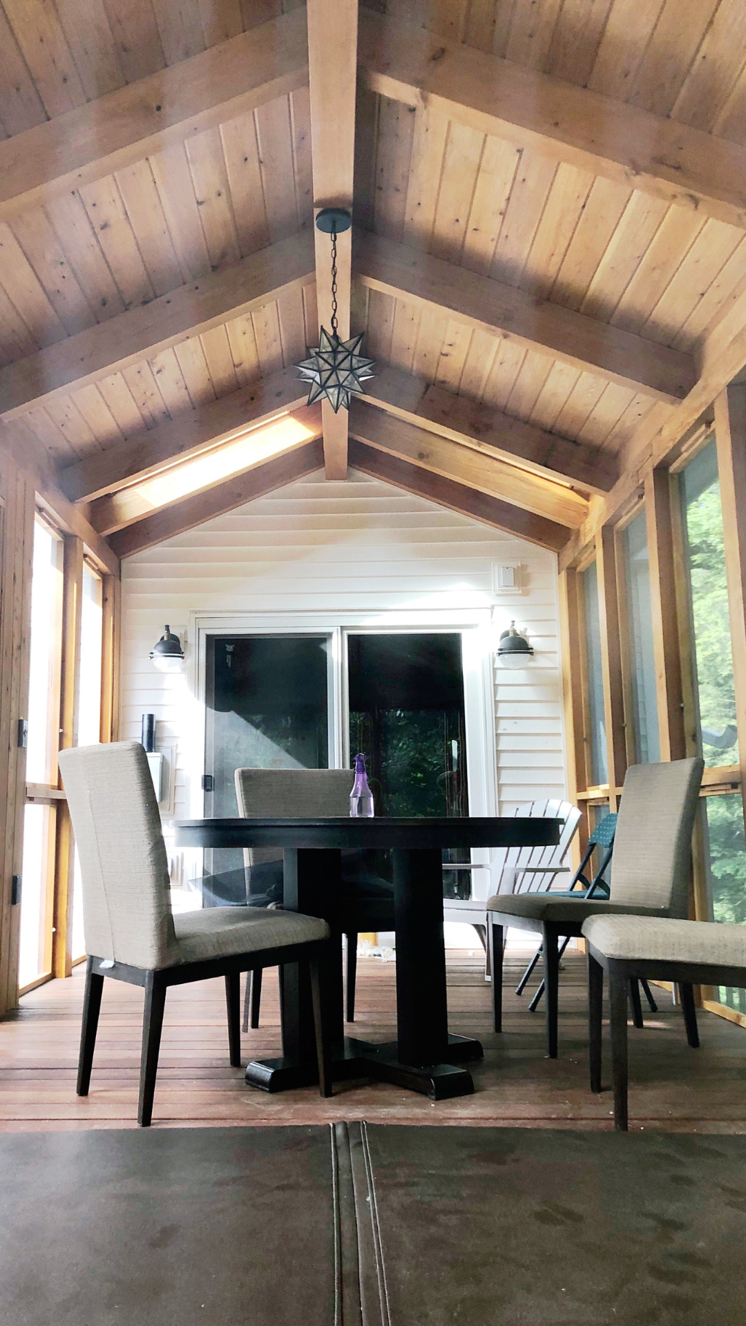 A screened porch with a wooden ceiling, a round black table, and six upholstered chairs. A bottle of spray cleaner is on the table. There are sliding glass doors and windows, with outdoor greenery visible through the windows.
