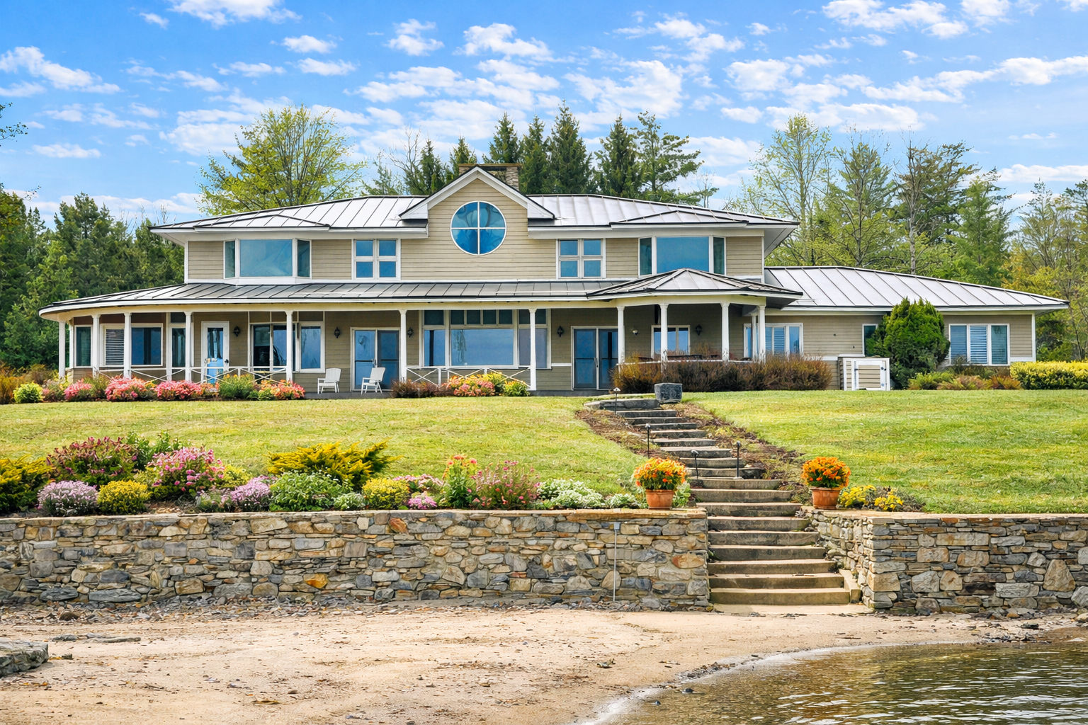 Large light gray house with a rounded window on the second floor, multiple rooflines, and a porch supported by white pillars, surrounded by a grassy yard with stone stairs leading up to it.