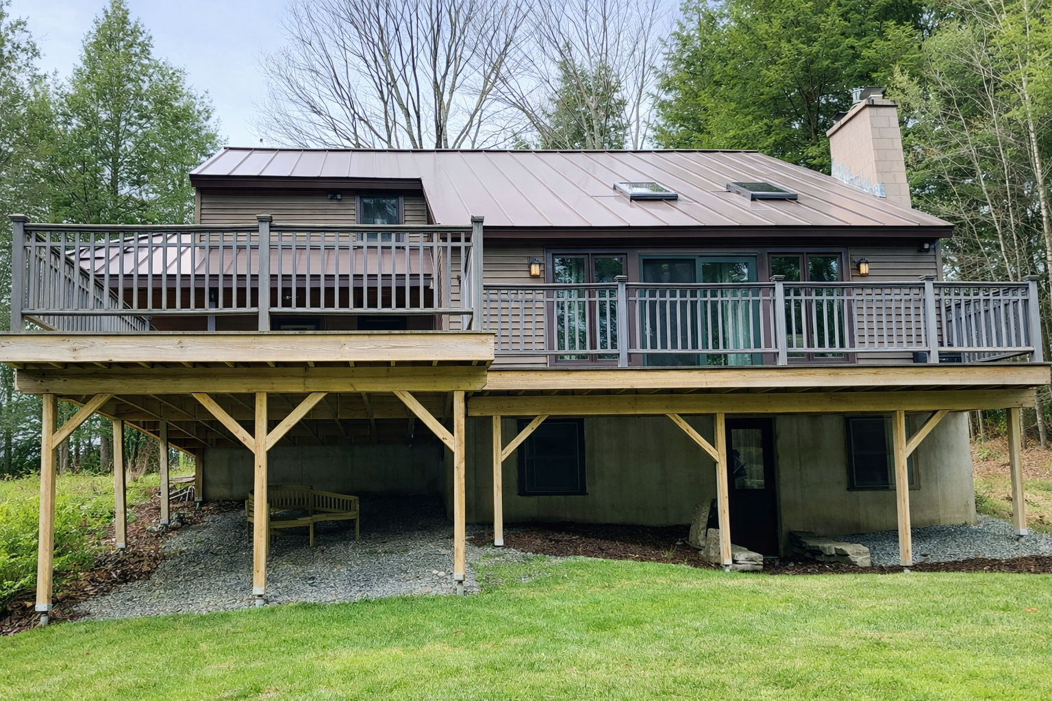 Back view of a two-story house with a wooden deck and railing, partially supported by wooden posts, with a grassy yard in the foreground and trees in the background.