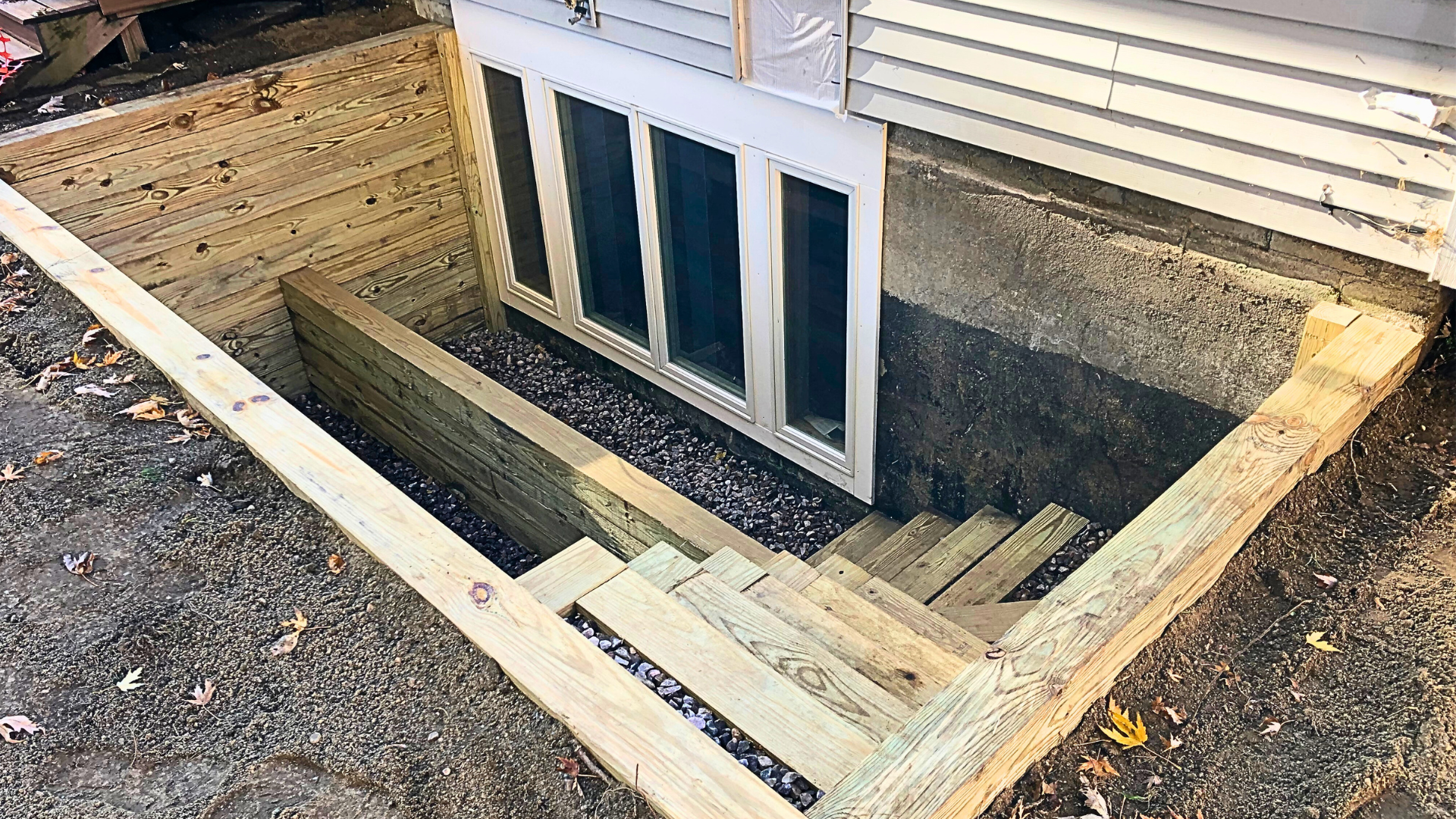 Construction of a wooden staircase and framing around a basement window with gravel ground and house siding.