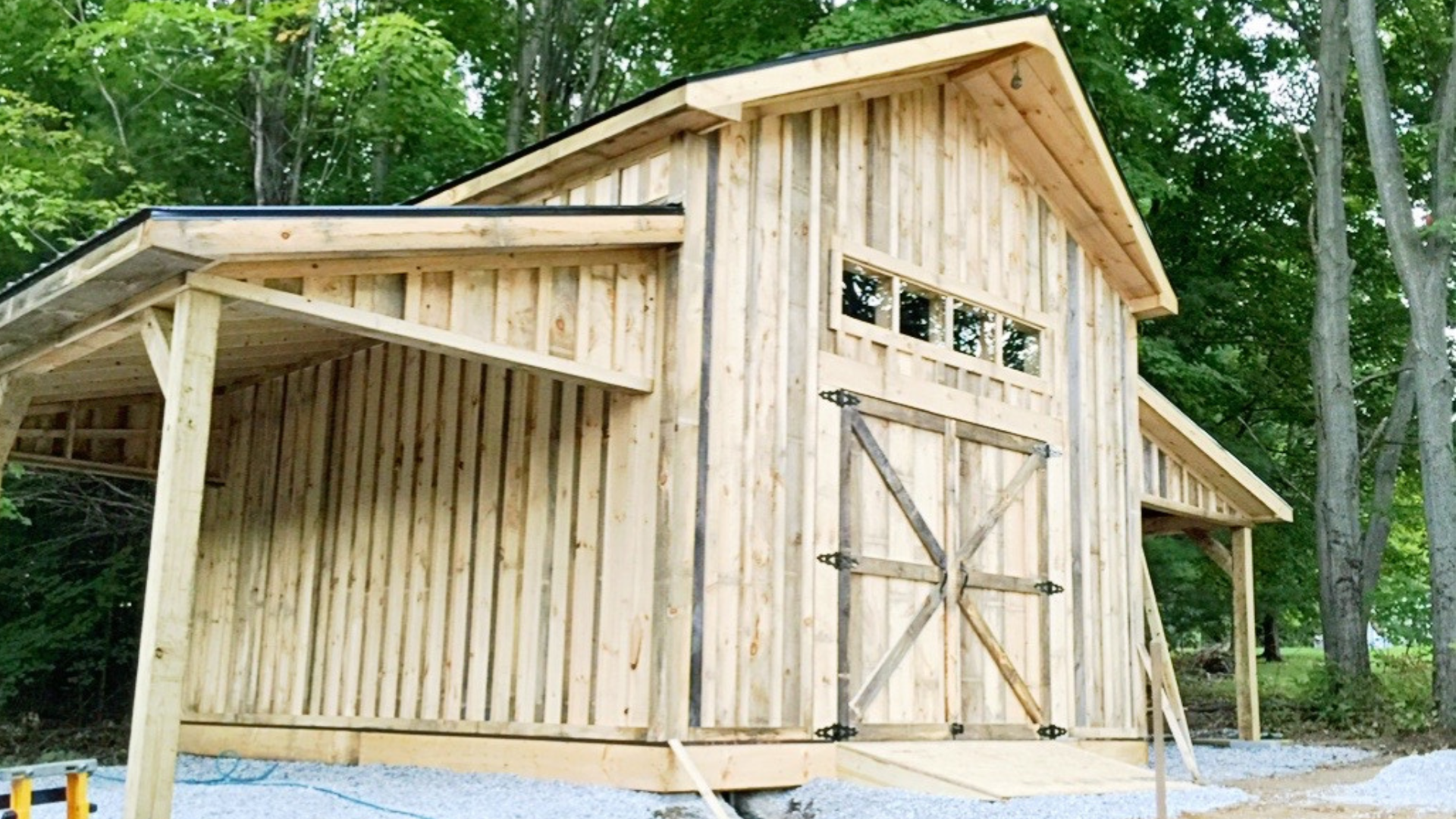 A wooden barn structure under construction in a wooded area, with a large door, small windows, and an overhanging roof.