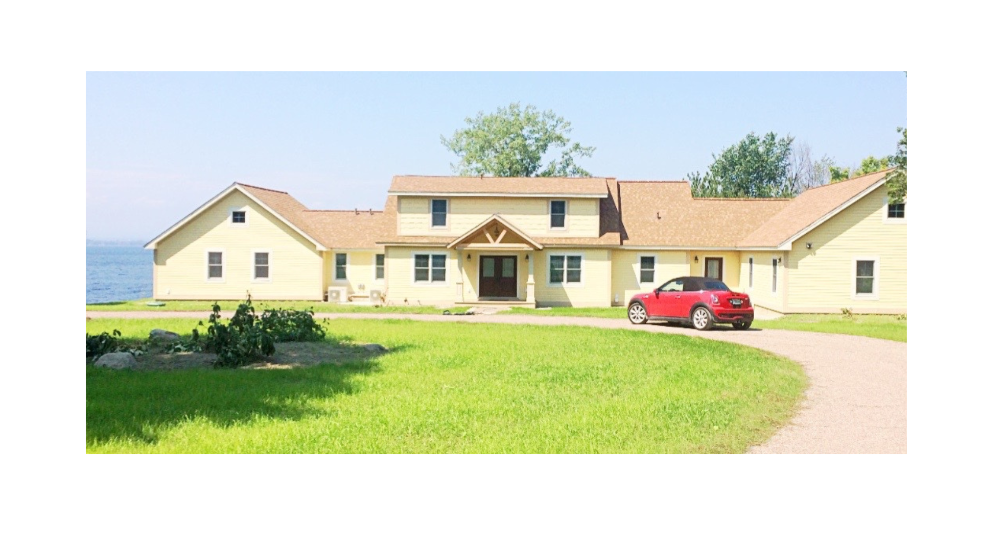 A large yellow house with a brown roof and attached garage, surrounded by a green lawn and a gravel driveway, with a red car parked in front, near a body of water under a clear blue sky.