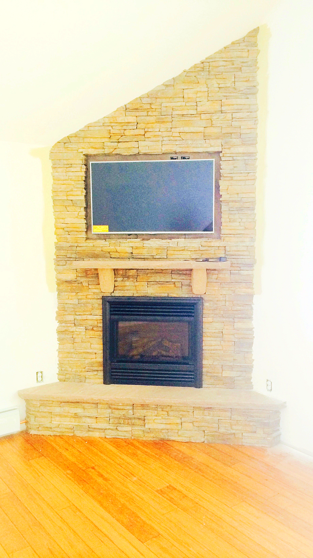 Living room with a stone fireplace and a mounted flat-screen TV above it, hardwood floors and white walls.