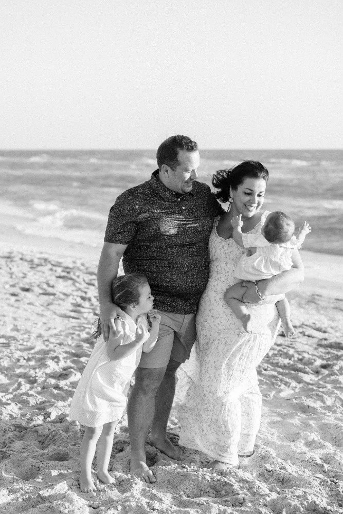 A family of four on a beach, includes a man, woman, and two young girls, smiling and enjoying time together.
