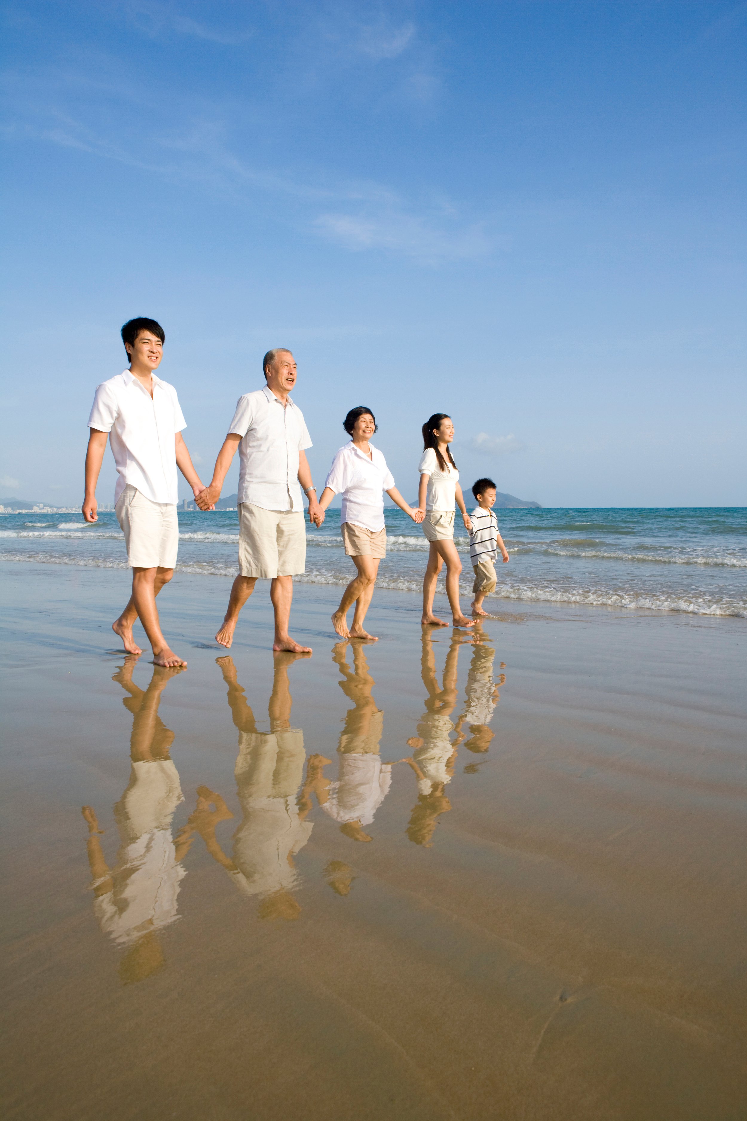 Family of six walking hand in hand on the beach, with their reflections visible in the wet sand, under a blue sky.
