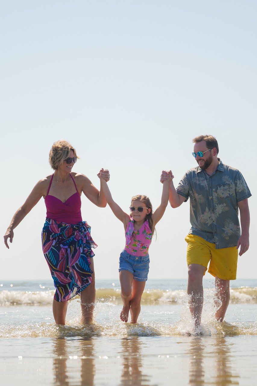 A family holding hands and walking in the shallow water at the beach on a sunny day, with the ocean in the background.