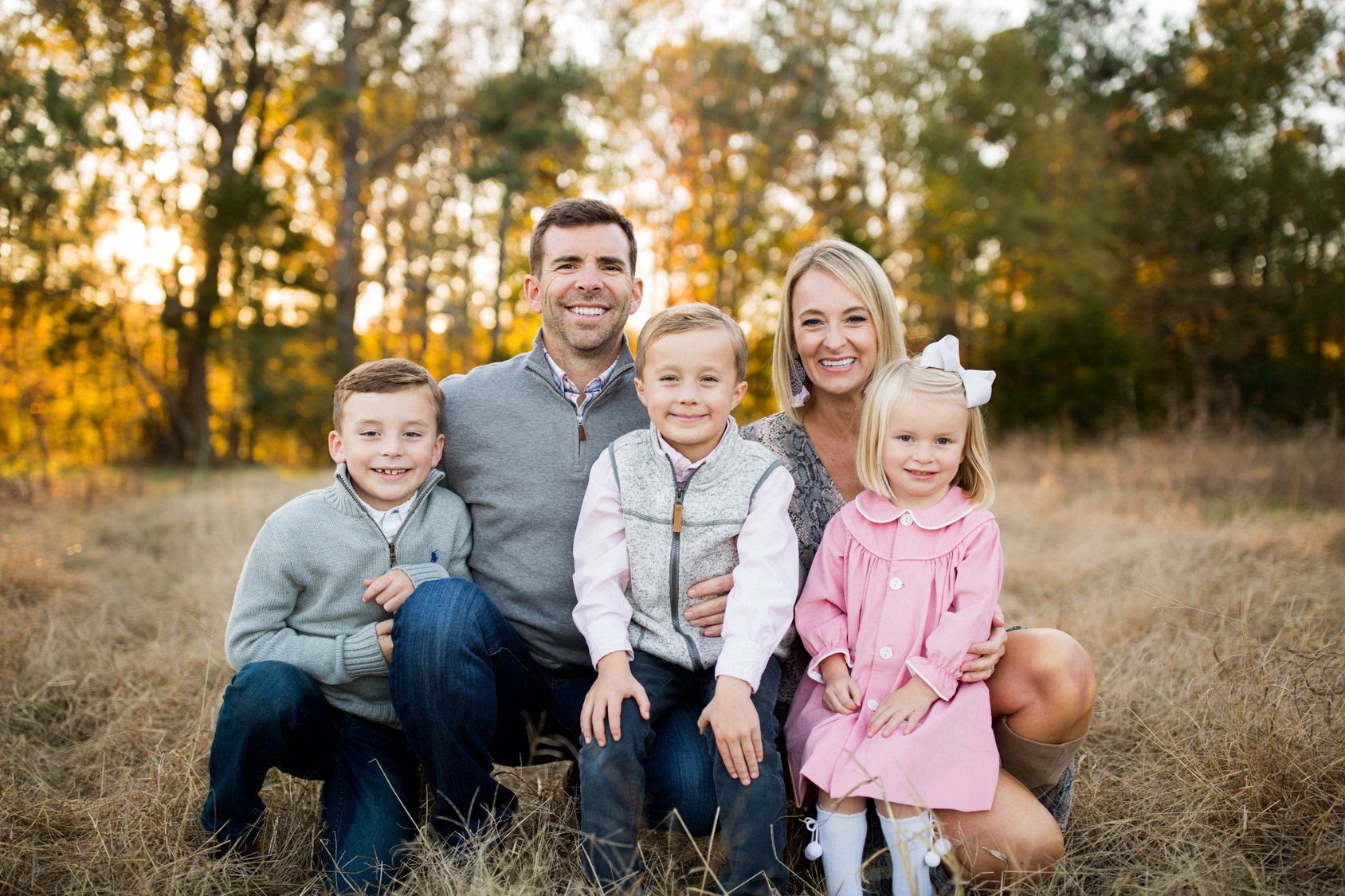A happy family of five posing outdoors in a field during autumn, with fall foliage in the background.