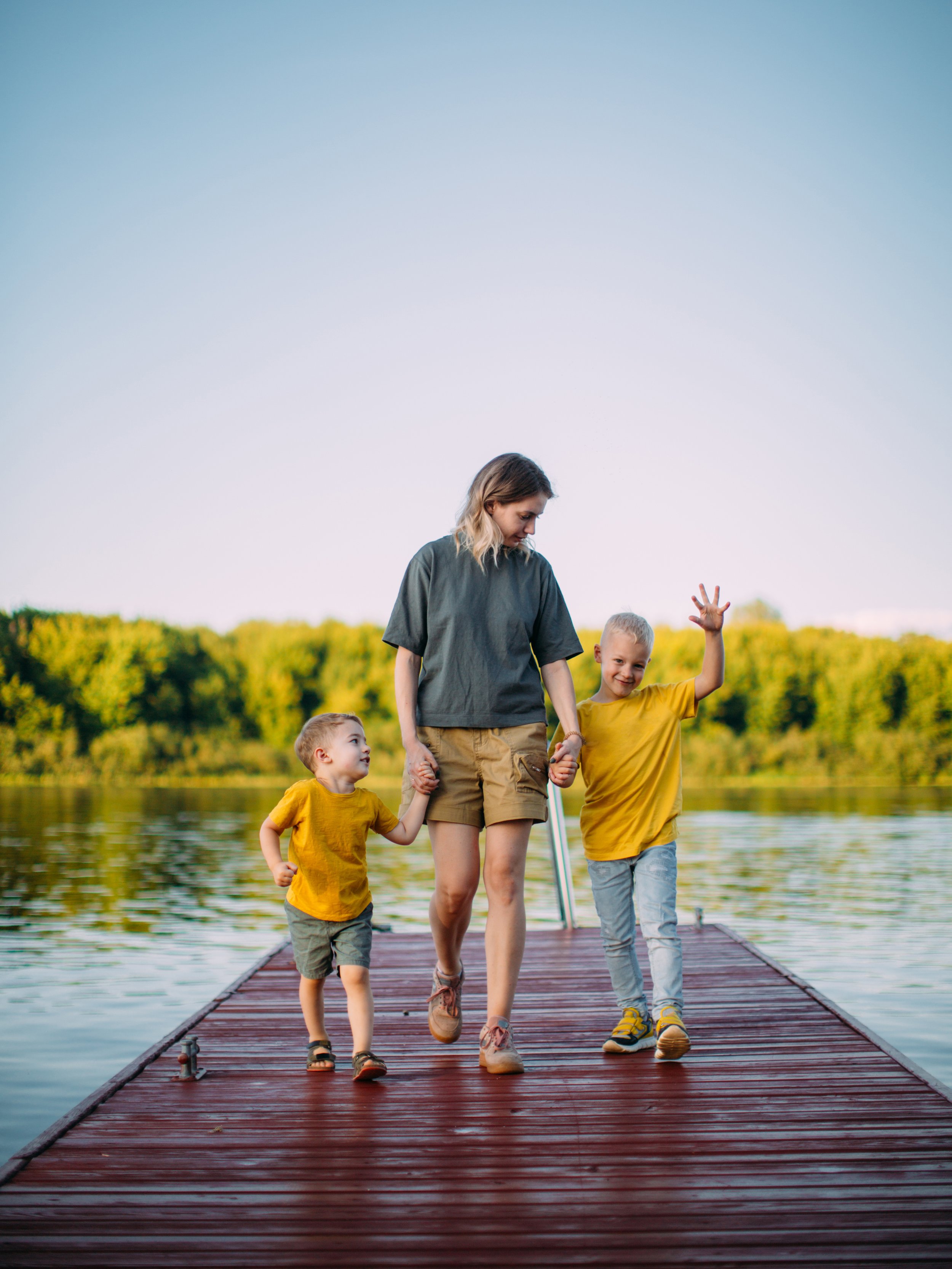 A woman walking on a wooden dock with two young boys by her side, holding hands and waving, near a calm lake with green trees in the background.