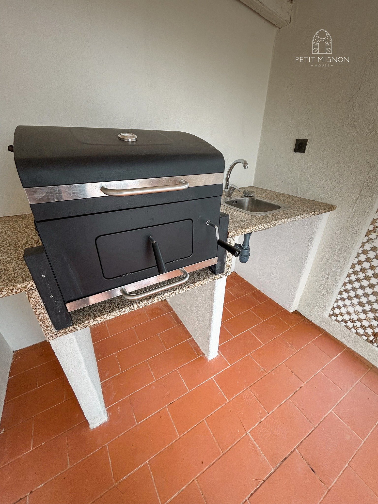 Churrasqueira elétrica preta sobre bancada de granito, próxima a pia de aço inox, em área de serviço com parede branca e piso de cerâmica vermelha.