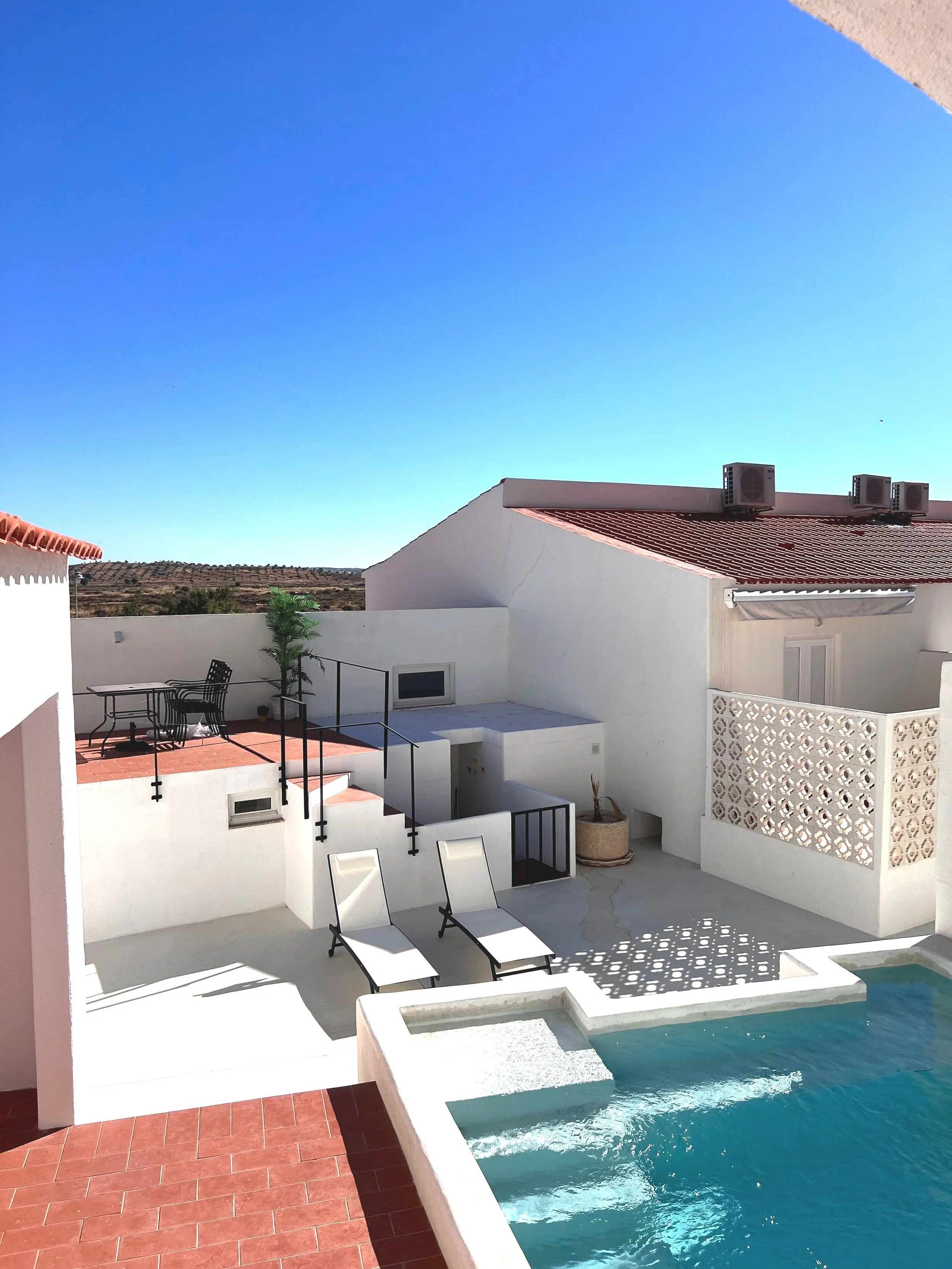 Vista de área da piscina ao ar livre com espreguiçadeiras brancas e sombra, fundo de casas brancas com telhados de barro e céu azul claro.