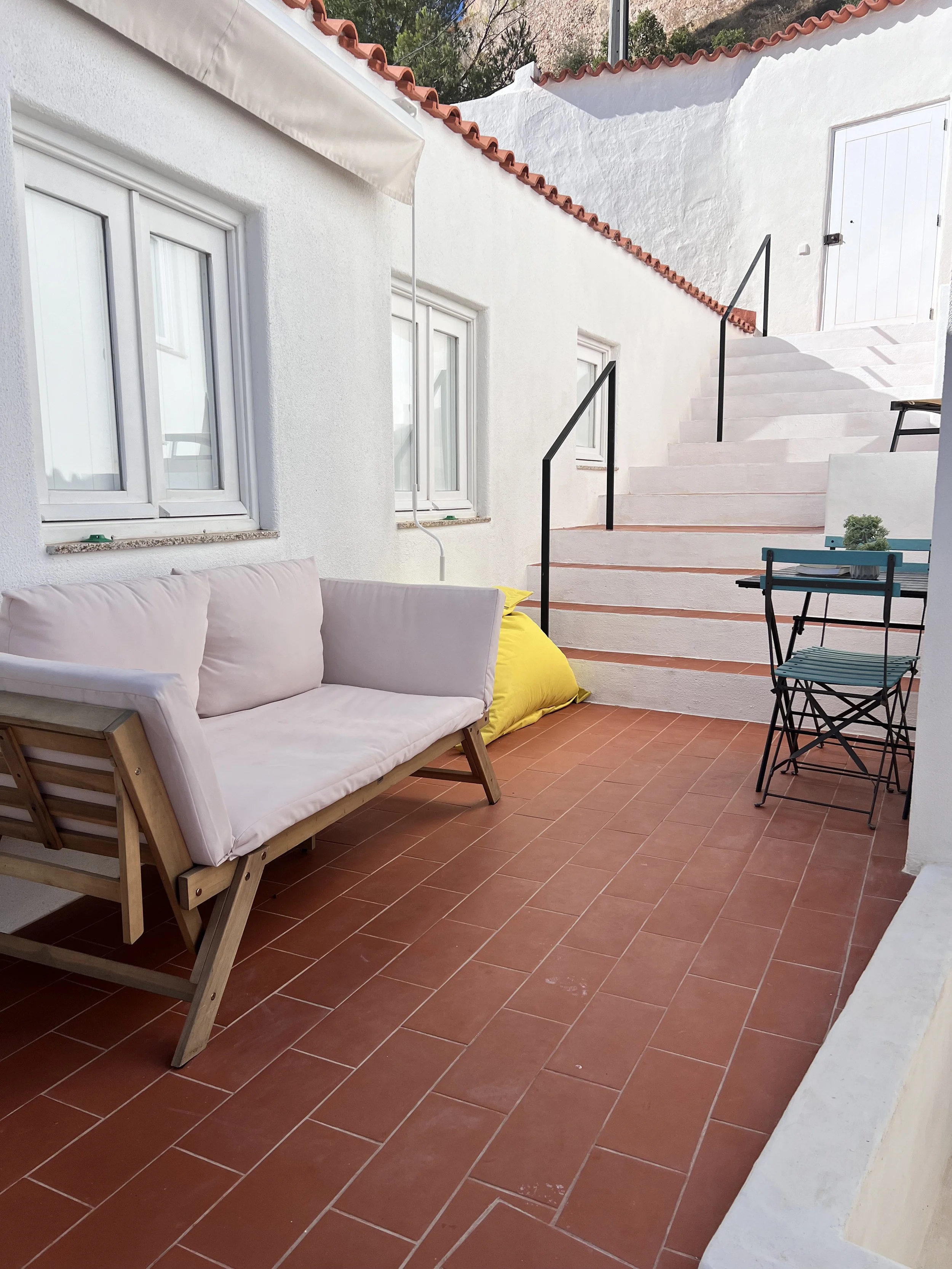 Varanda com sofá de madeira com almofadas brancas, uma mesa pequena, uma rua de degraus brancos com corrimão de barra preta, duas janelas brancas na parede e uma porta de correr branca.