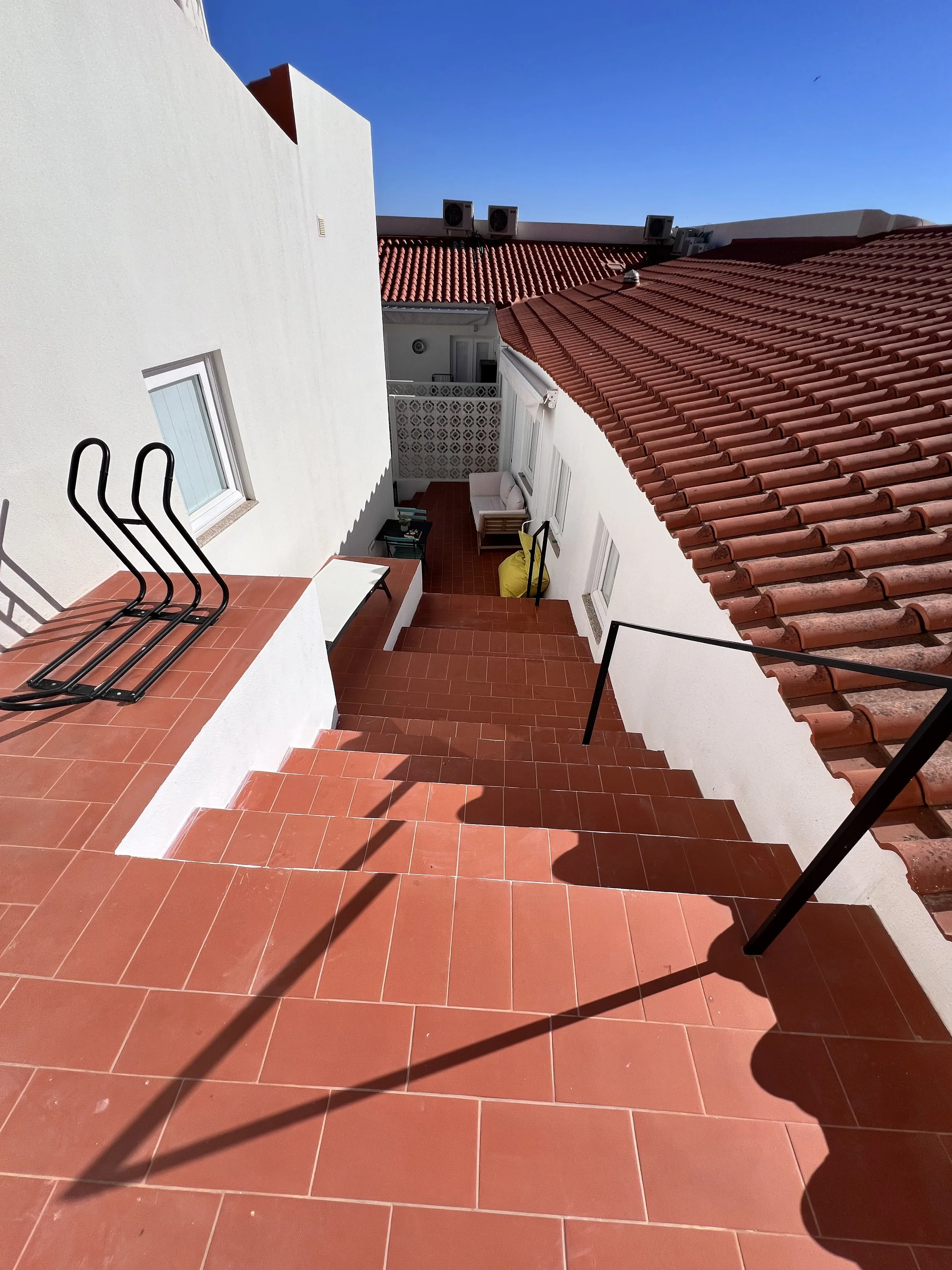 Escada de degraus de terraço com tubos de suporte de ferro, paredes brancas, telhado de telhas vermelhas, área de estar ao fundo com sofá, almofada amarela, e céu claro ao fundo.