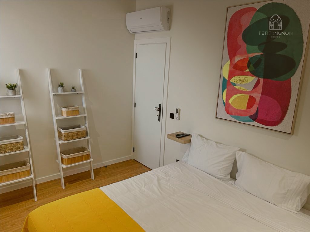 Quarto com cama de casal com lençóis brancos e travesseiros, uma parede com arte abstrata colorida, duas prateleiras brancas com plantas e cestas, uma porta branca, um ar-condicionado no alto, e um pequeno móvel com controle remoto ao lado da cama.