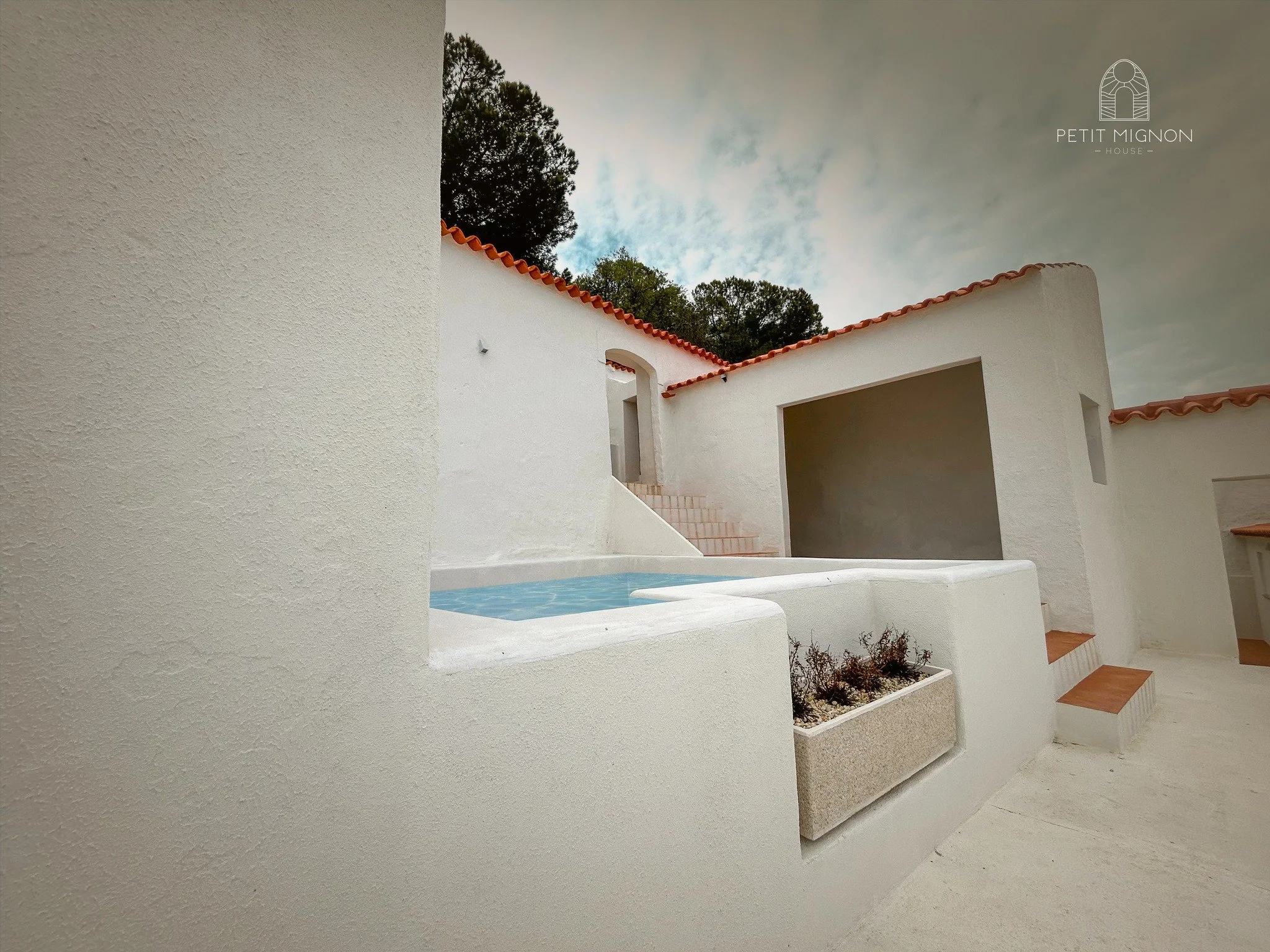 Casa de pátio branco com uma piscina pequena e escadas, teto com telhas de cerâmica, árvores ao fundo e céu nublado.