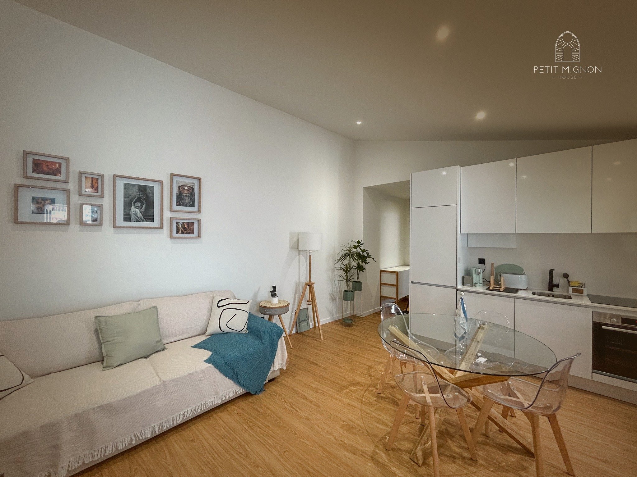 Sala de estar com sofá bege, quadros na parede, luminária, mesa de jantar de vidro com cadeiras transparentes, cozinha branca ao fundo, piso de madeira.