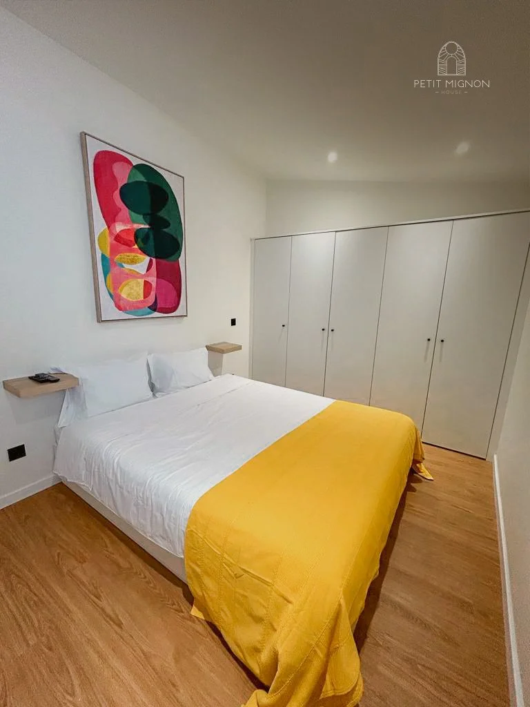 Quarto de hotel com cama de casal com colcha amarela, paredes brancas, armário embutido branco, quadro com arte abstrata com cores vibrantes na parede, piso de madeira e duas pequenas prateleiras ao lado da cama.