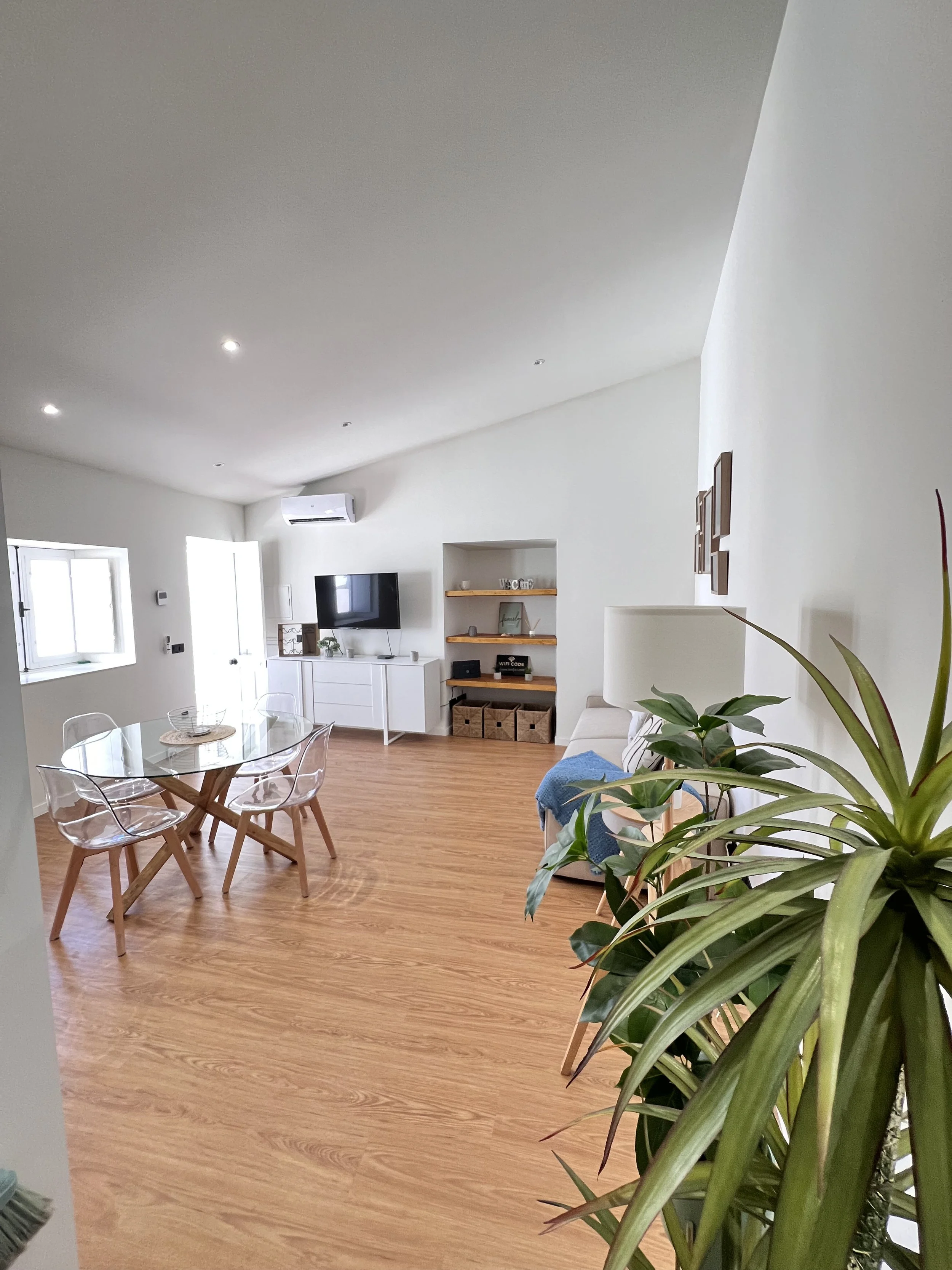 Sala de estar moderna com móveis brancos, televisão na parede, piso de madeira, janelas grandes, plantas e iluminação embutida no teto.