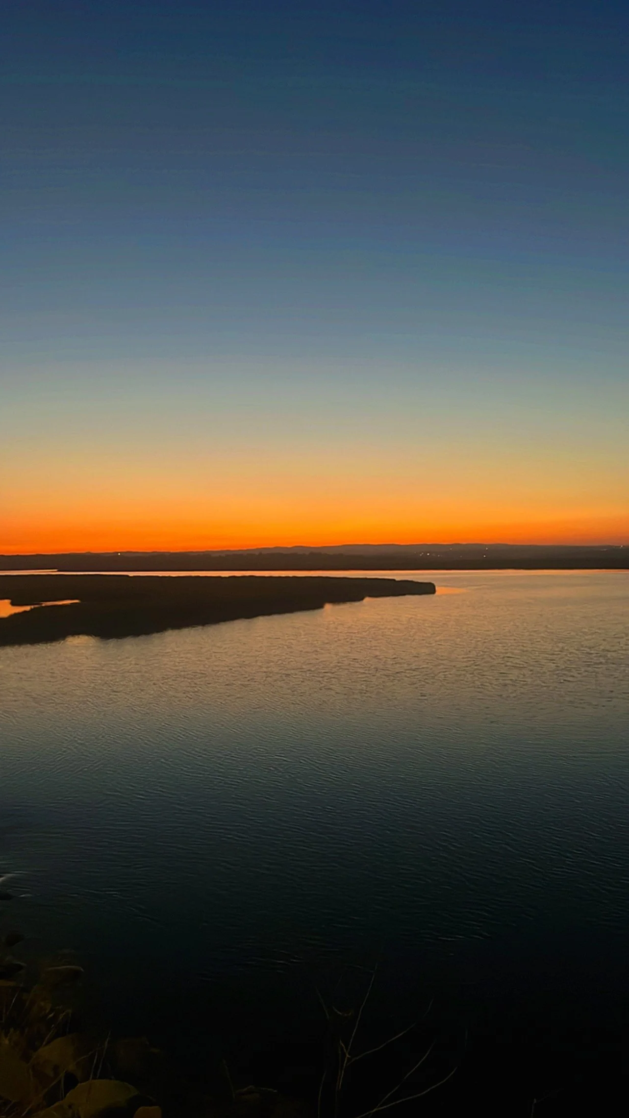 Céu ao pôr do sol acima de um rio, com tons de laranja, amarelo, azul e cinza, refletidos na água.